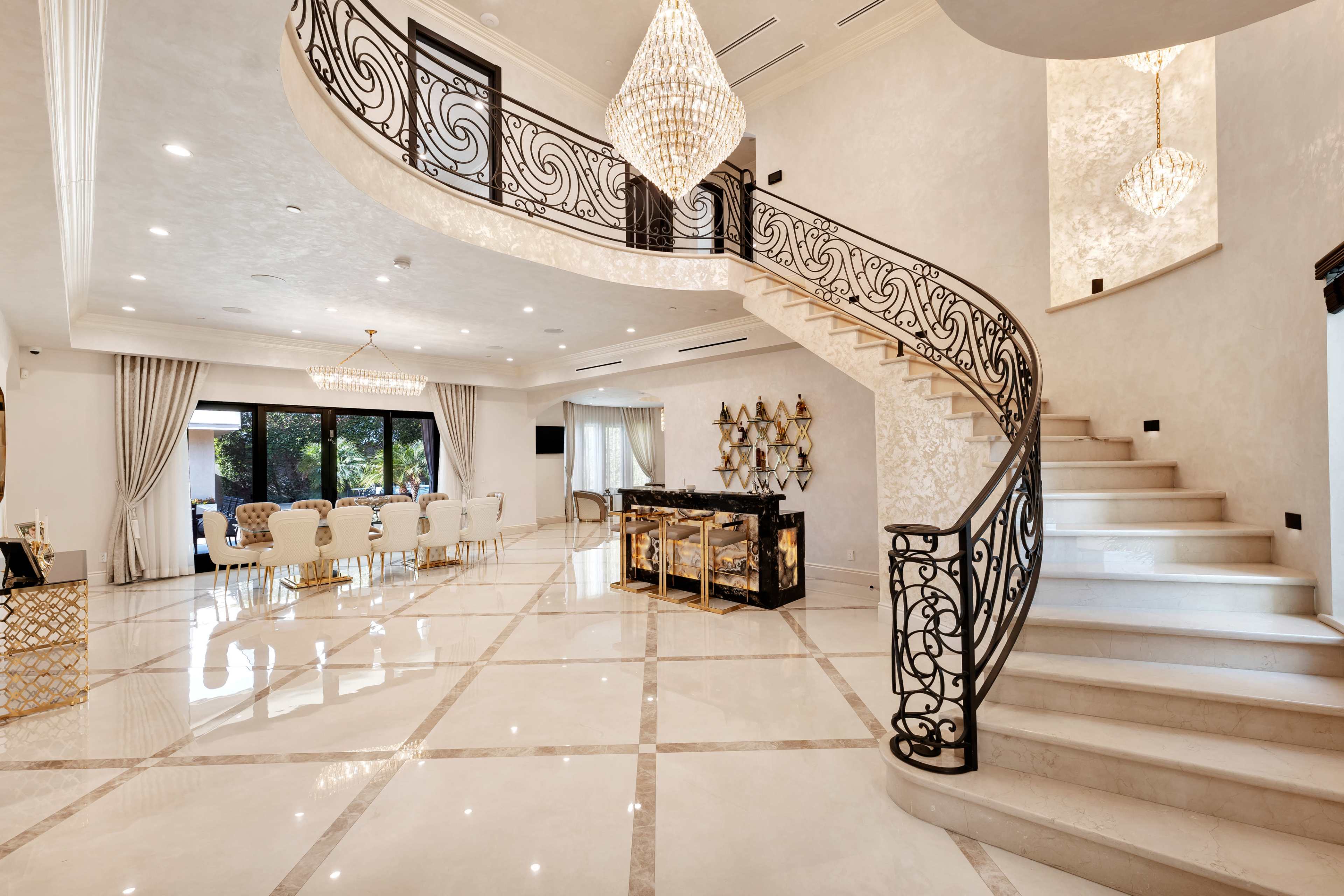 The image shows a luxurious interior with a sweeping staircase, ornate iron railings, and a spacious dining area with elegant furniture.
