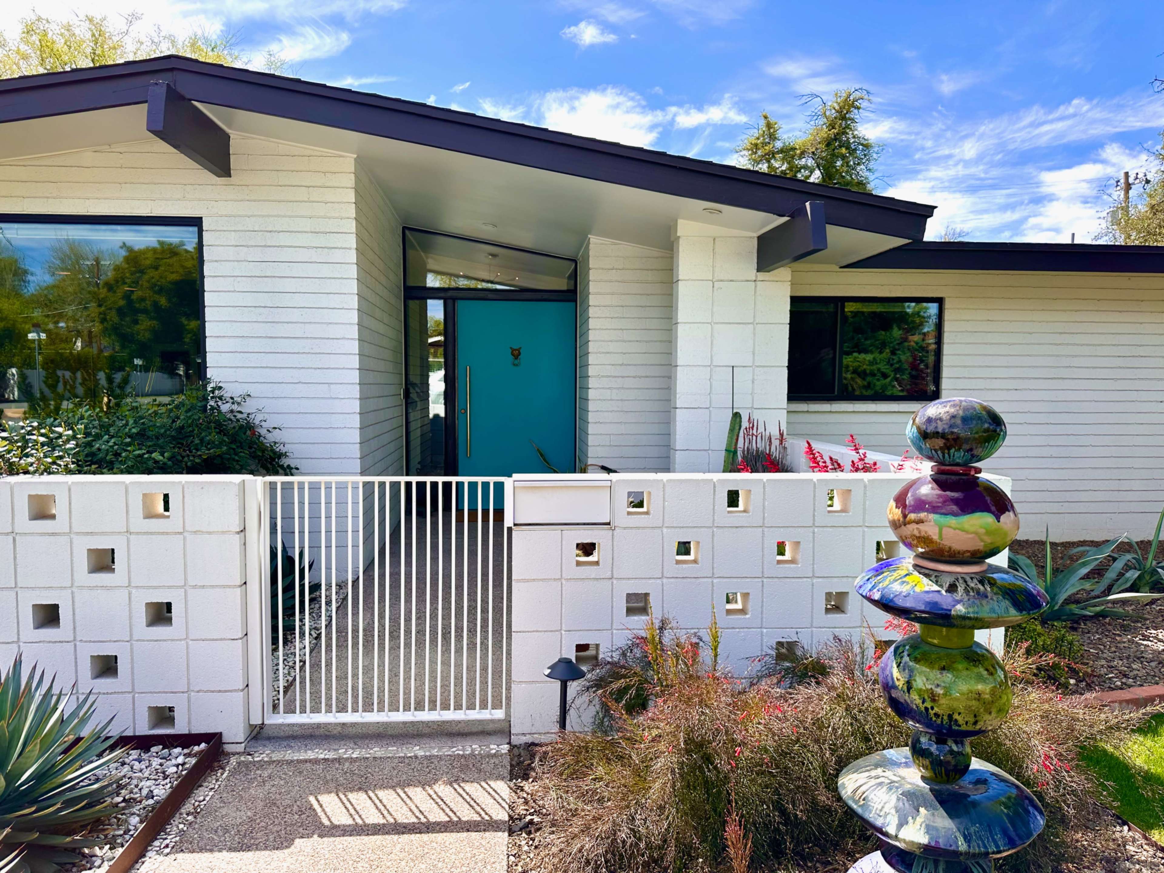 A modern house features a turquoise door, a gated entrance with decorative blocks, and a colorful sculpture in the yard.