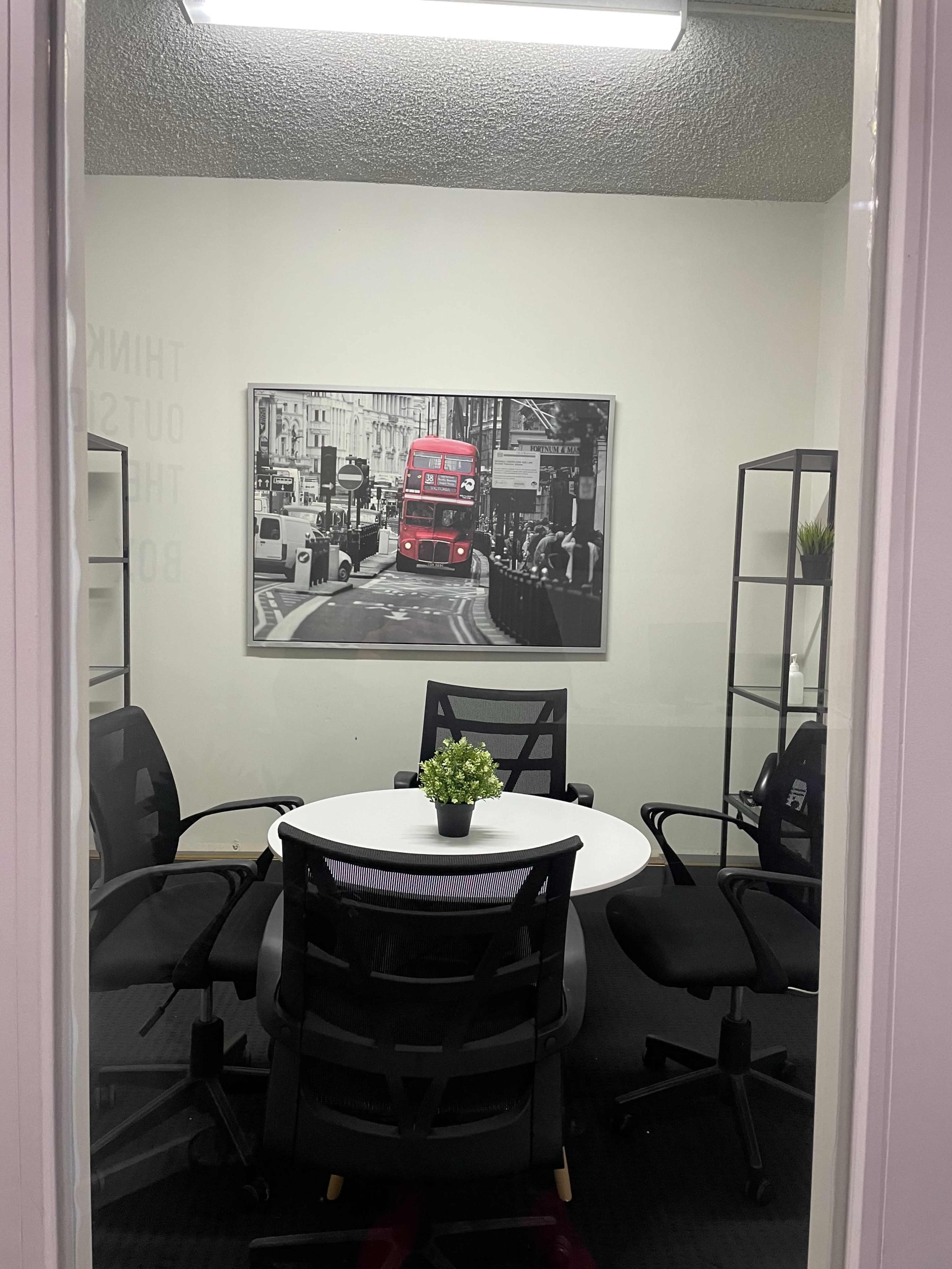 A round table with black chairs is set up in a small conference room, featuring a framed black-and-white photo of a red double-decker bus on the wall.