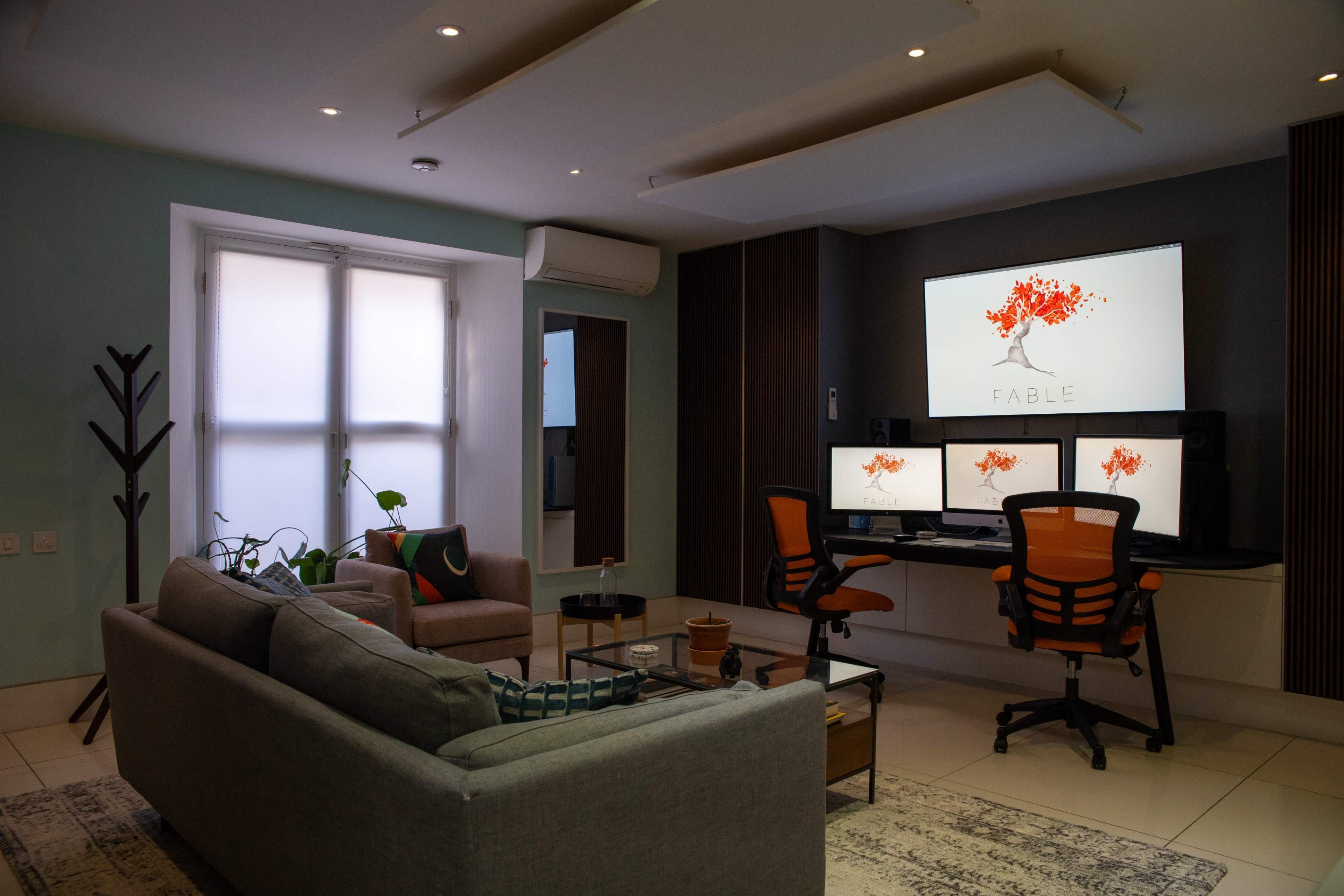 The image shows a modern workspace with a comfortable seating area, featuring a large wall-mounted screen displaying a logo for "FABLE," and multiple computer monitors on a desk.