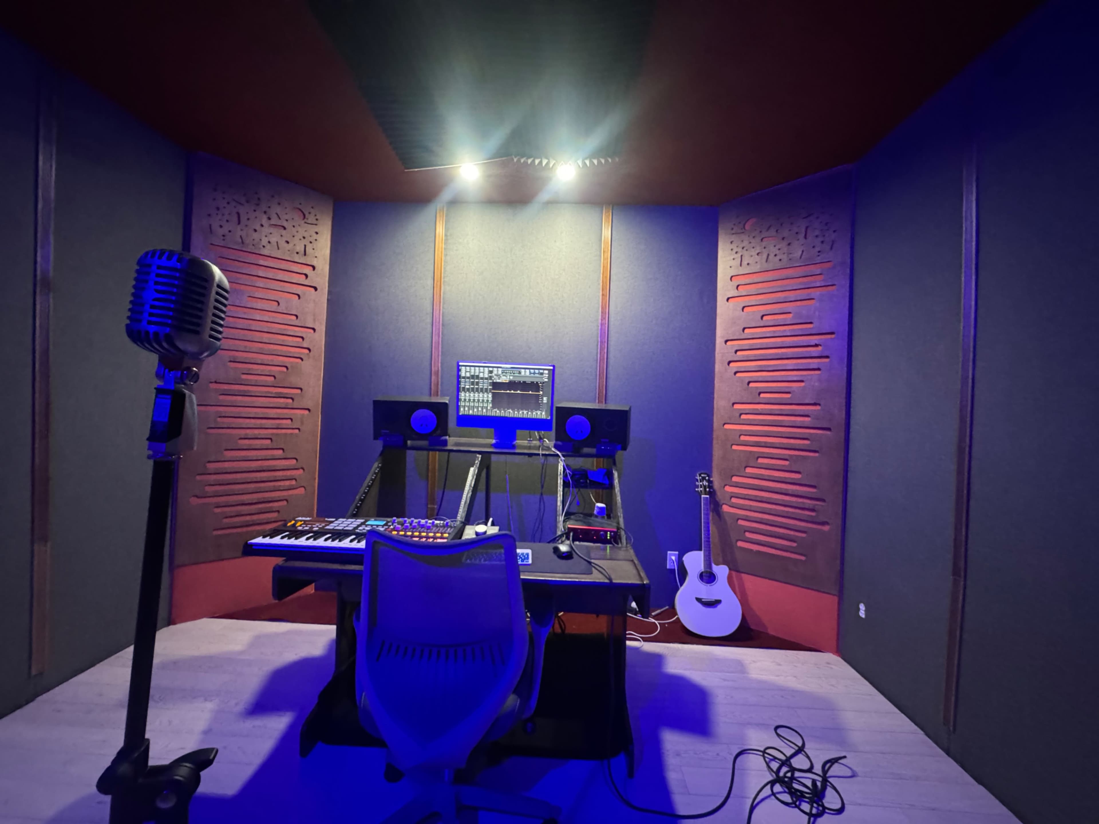 The image shows a music studio with a central desk containing a keyboard and audio equipment, flanked by soundproofing panels and a microphone stand.