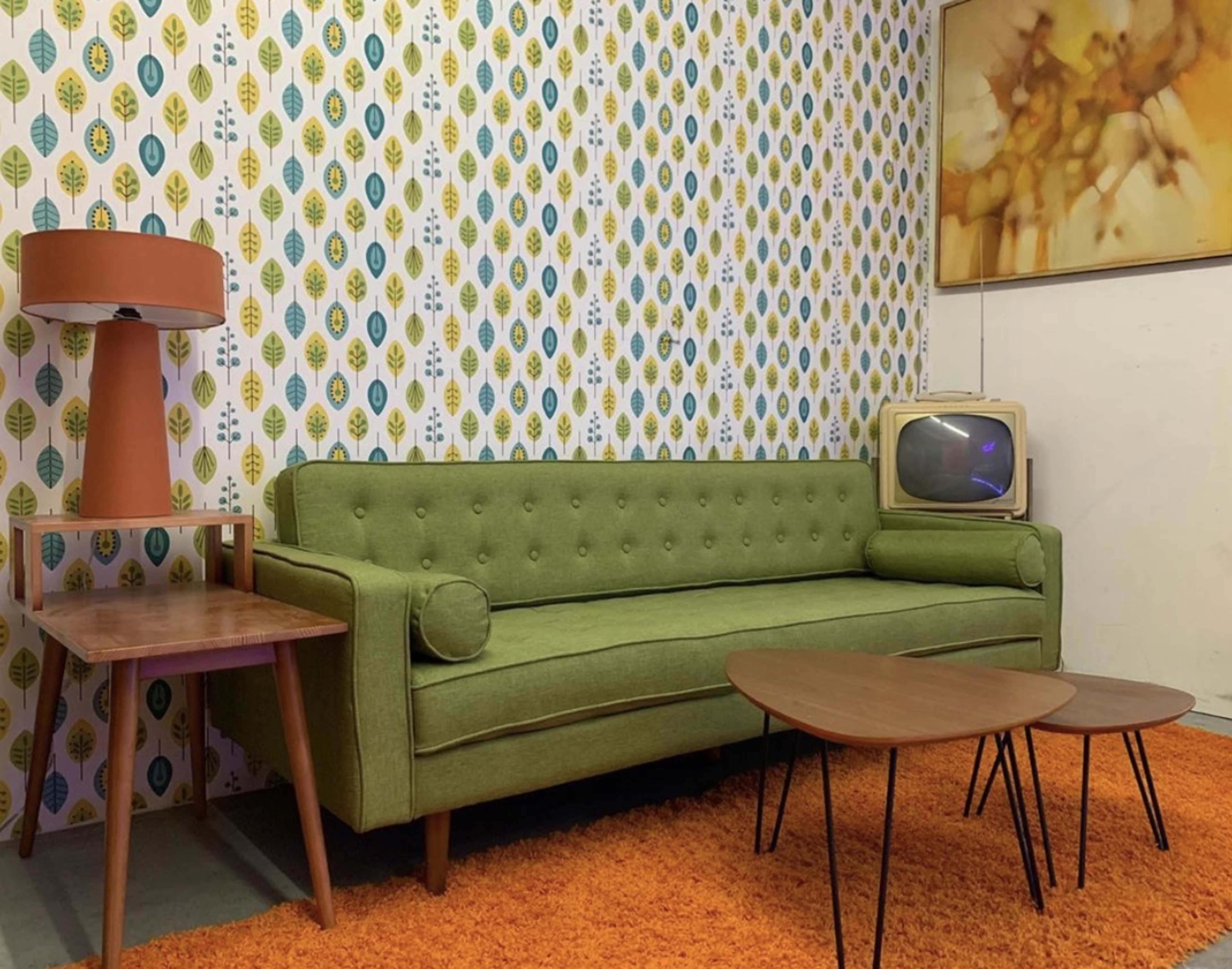 The image shows a vintage living room with a green sofa, a side table, a retro lamp, and a small television set against a patterned wallpaper.