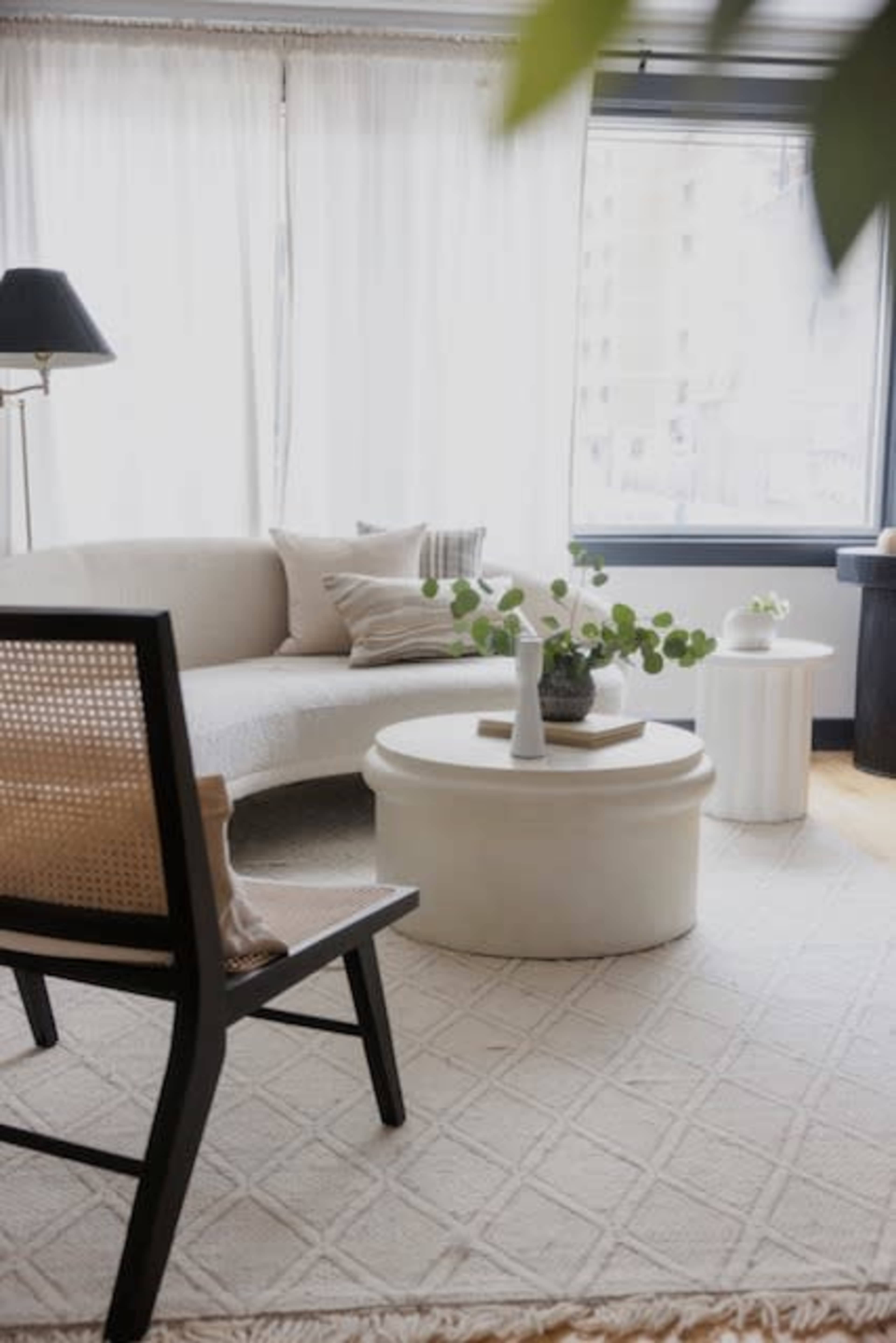 A modern living room with a curved sofa, a round coffee table, and a chair, all set against large windows draped with sheer curtains.