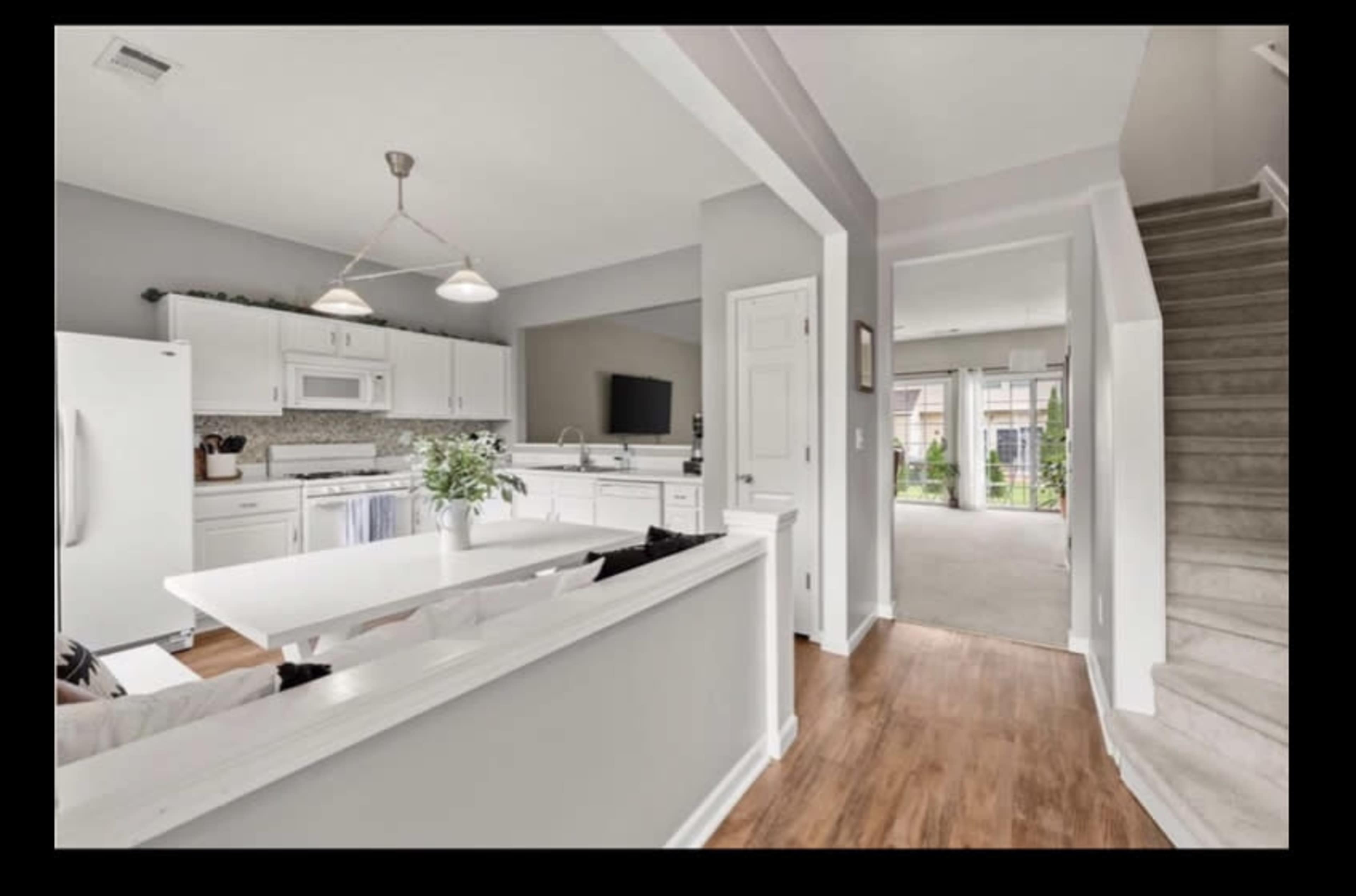 The image shows a modern kitchen with white cabinets and appliances, connected to a dining area and visible staircase, leading to a carpeted living space.
