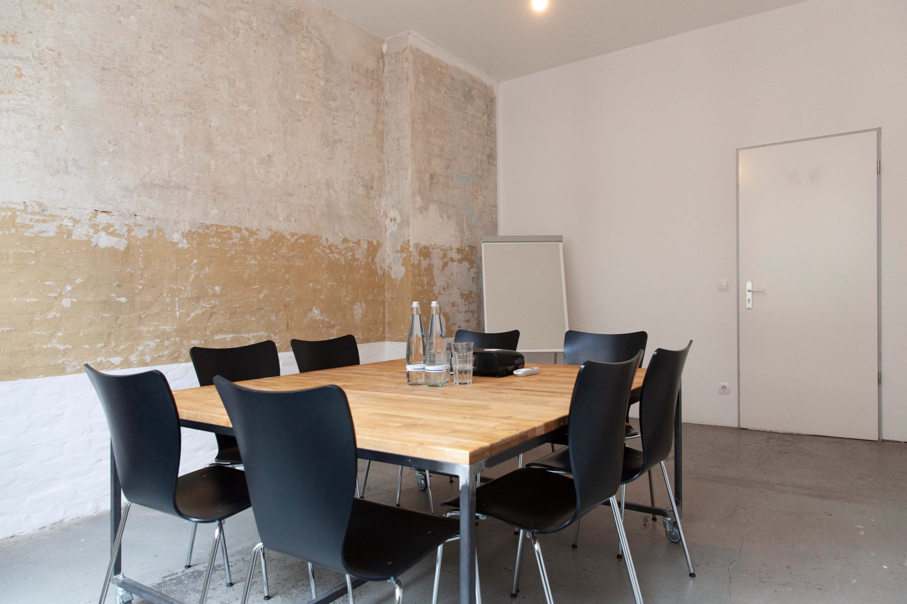 A conference room features a wooden table surrounded by black chairs, with a whiteboard and glass of water on the table.