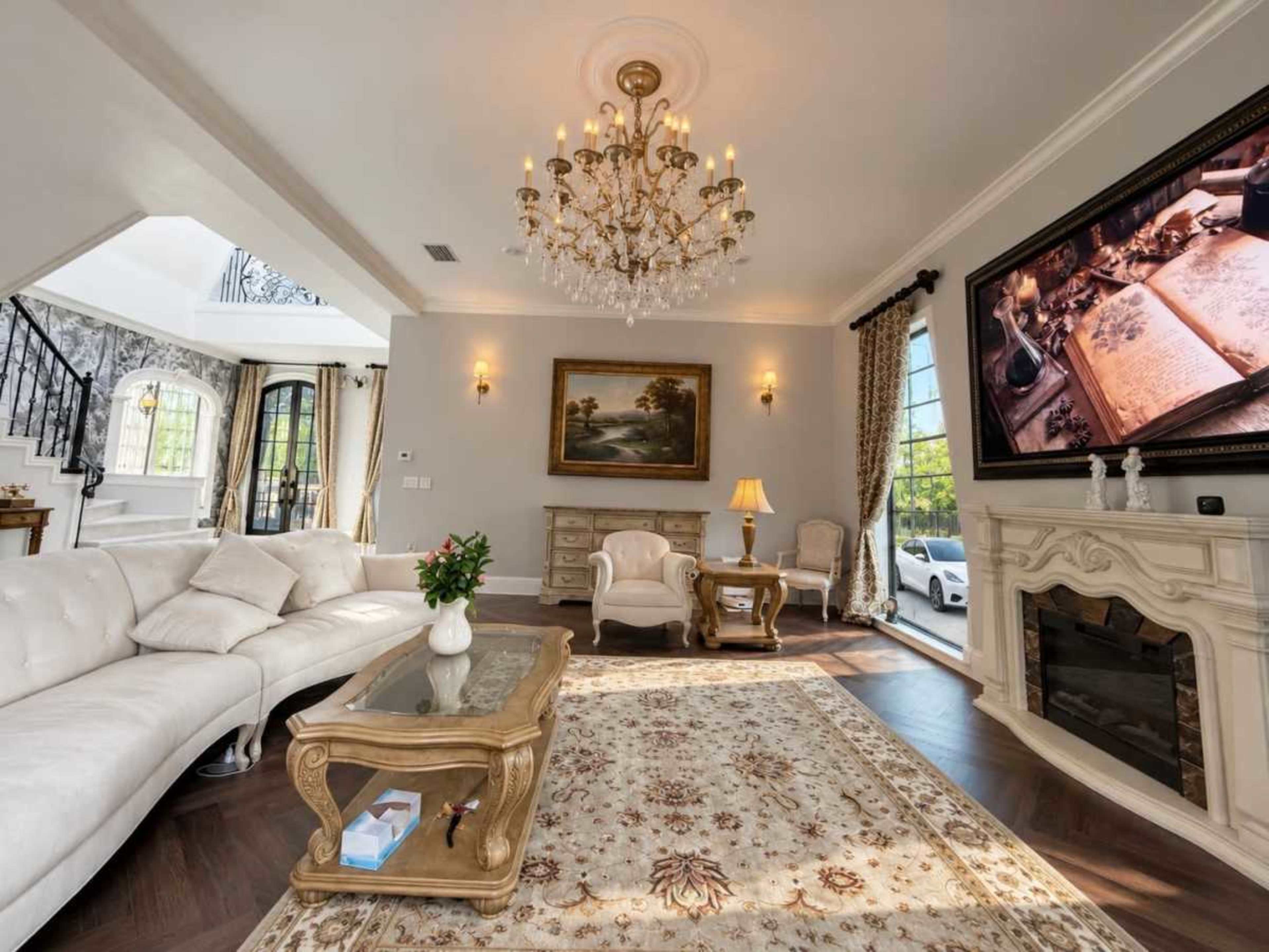 The image shows a spacious living room featuring a large white sectional sofa, an ornate chandelier, and a decorative painting, with a fireplace and a flat-screen TV mounted on the wall.