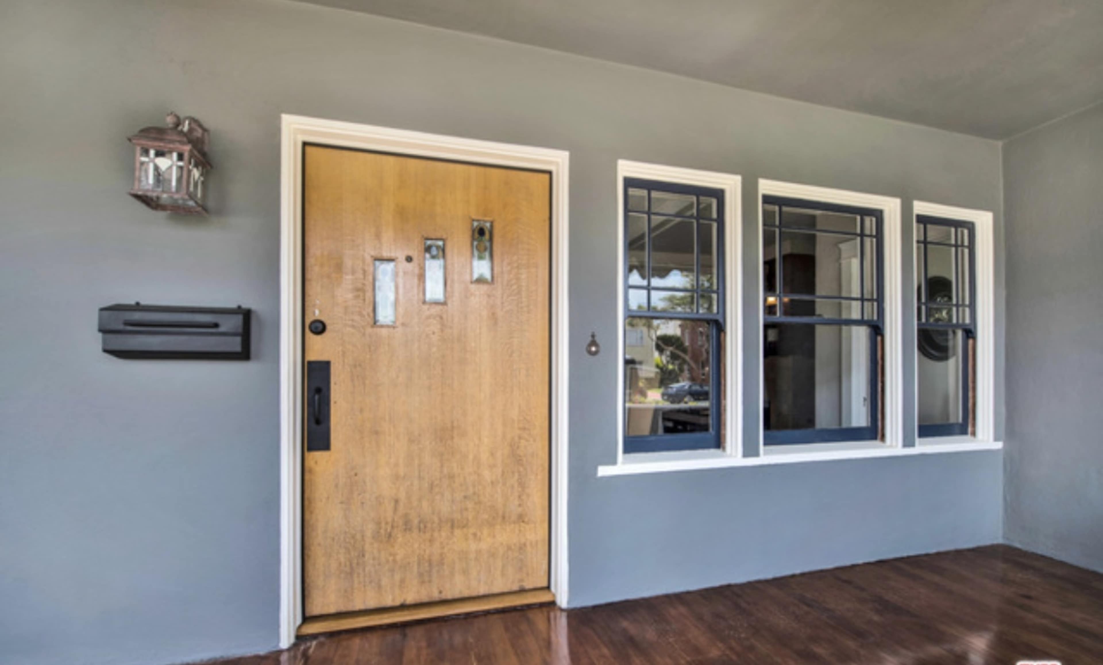The image shows a front porch with a wooden door featuring vertical glass panels, flanked by two windows, and a lantern beside the entrance.