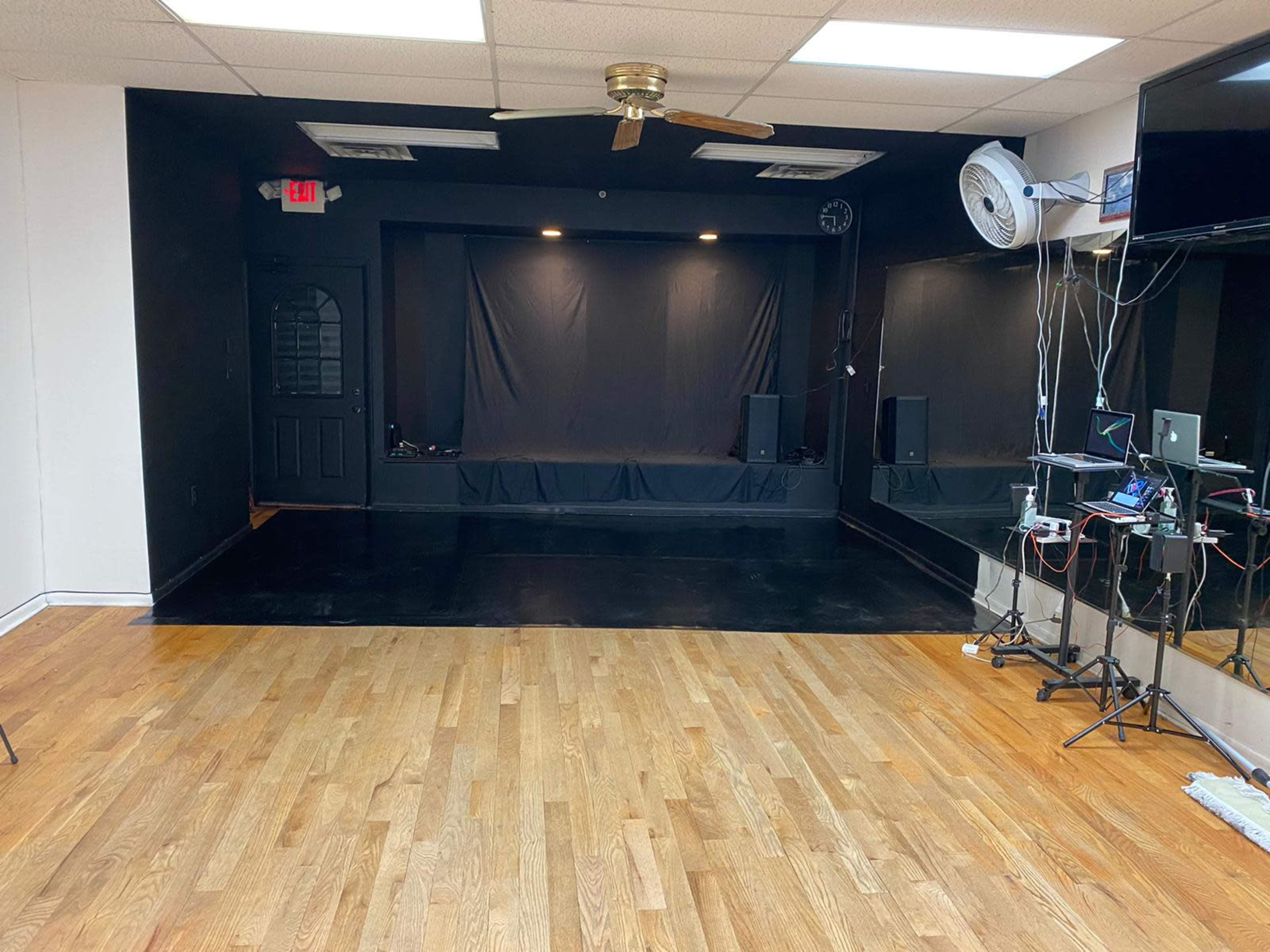 A spacious empty performance room with black curtains, a wooden floor, and music equipment set up on one side.