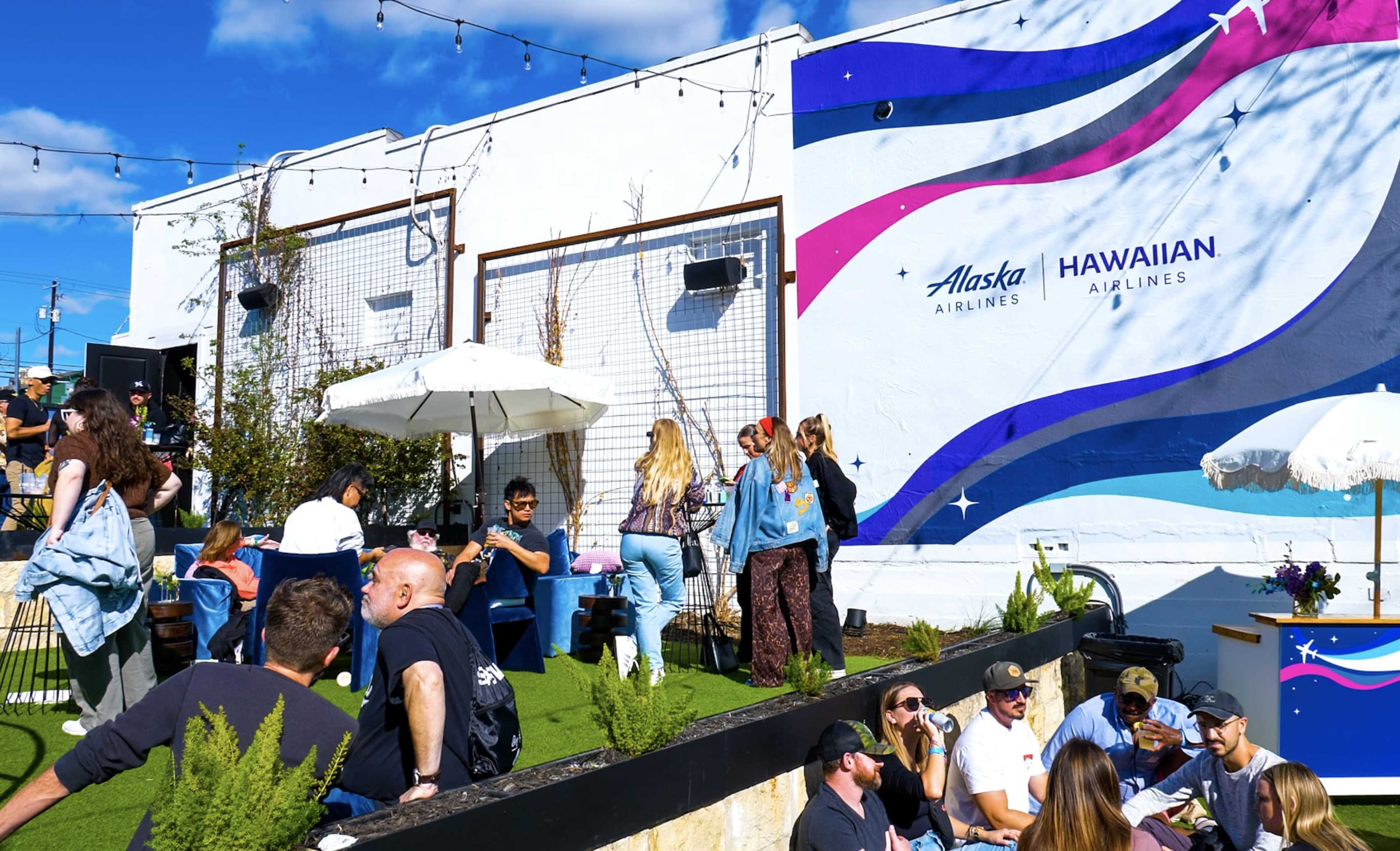 A group of people enjoy a social gathering in an outdoor lounge area decorated with a mural featuring Alaska Airlines and Hawaiian Airlines branding.