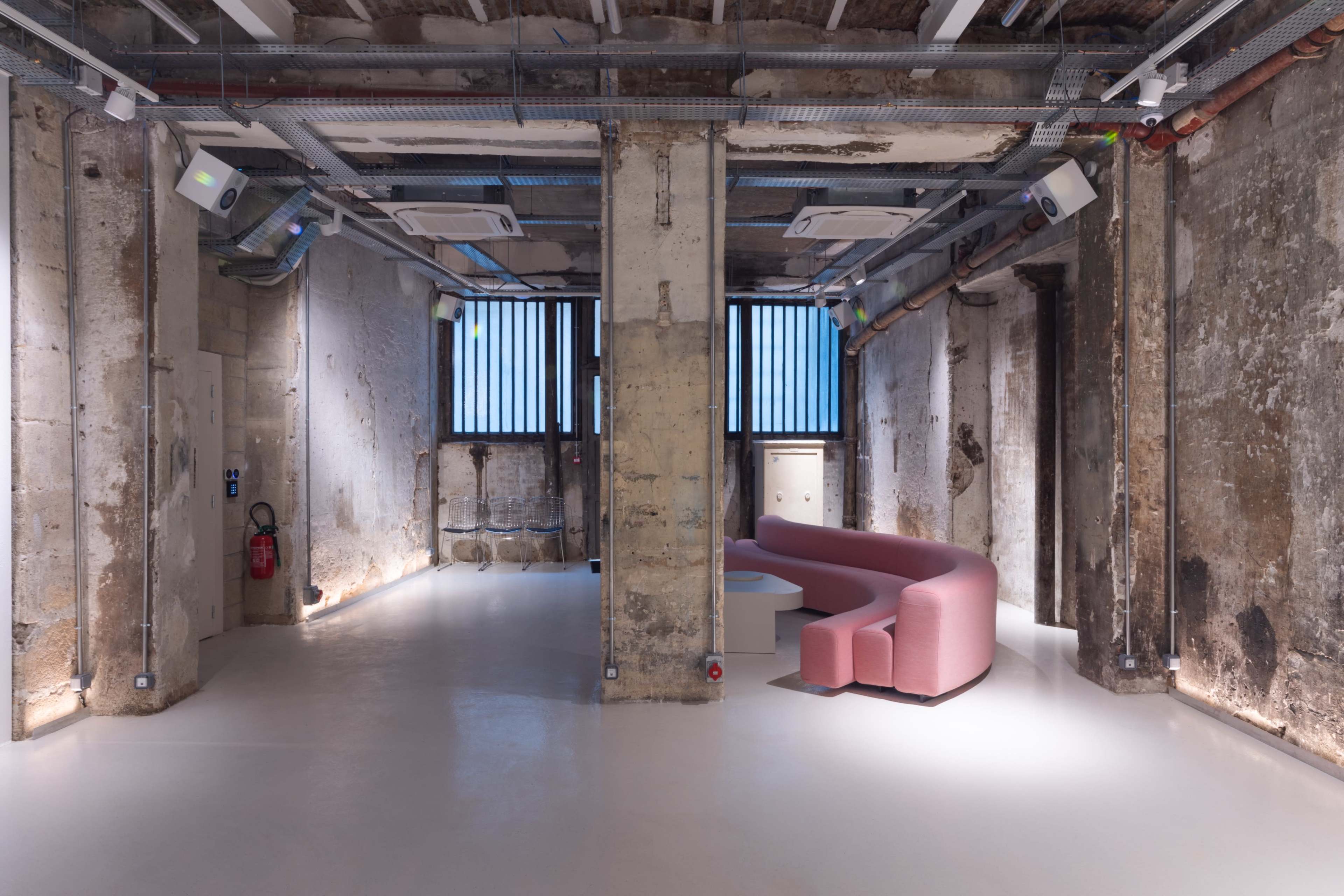 The image shows a spacious industrial-style interior with exposed concrete pillars and a pink curved sofa in the center.
