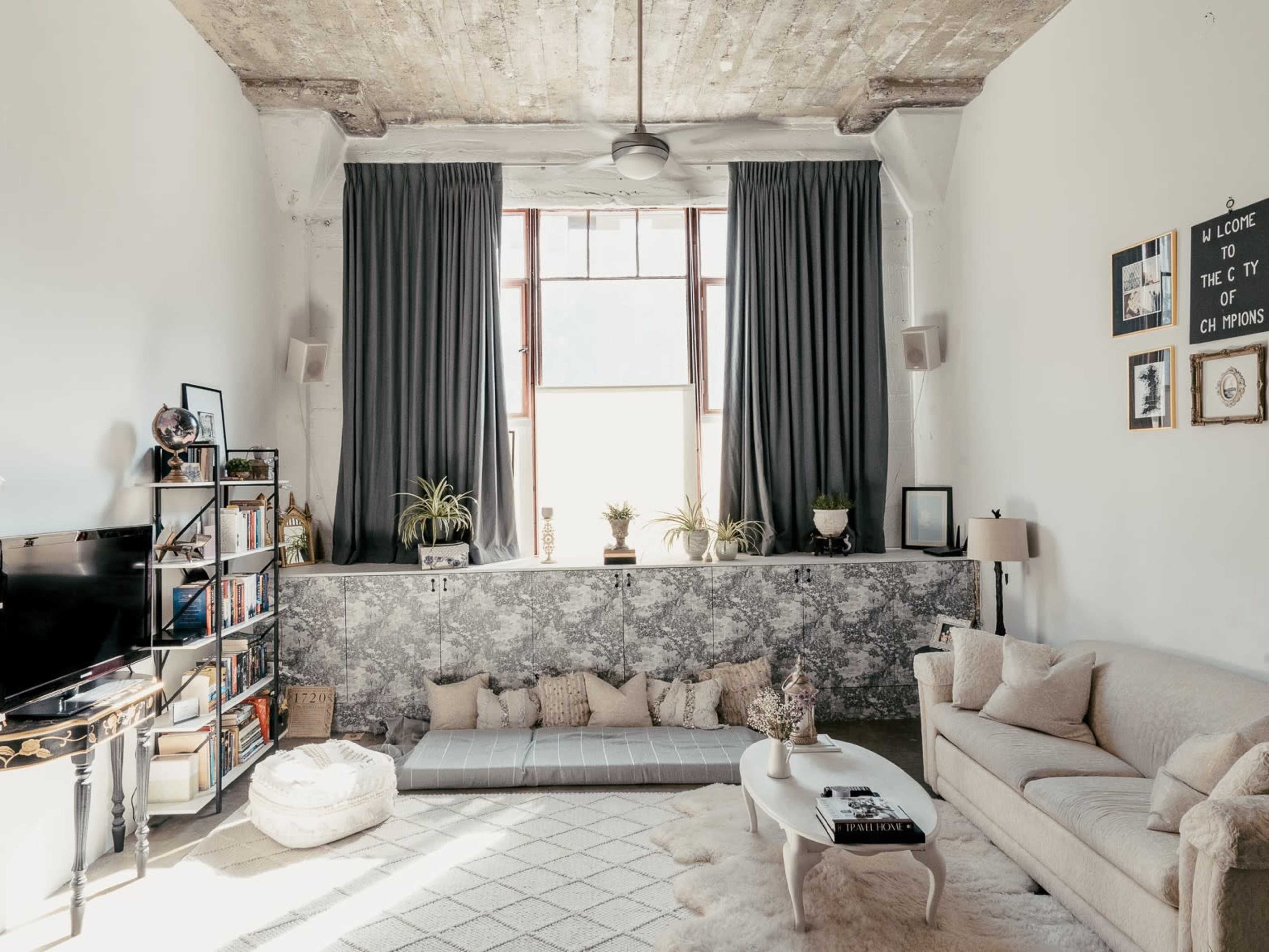 The image features a spacious living room with large windows, a low seating area, a light-colored sofa, and decorative plants.