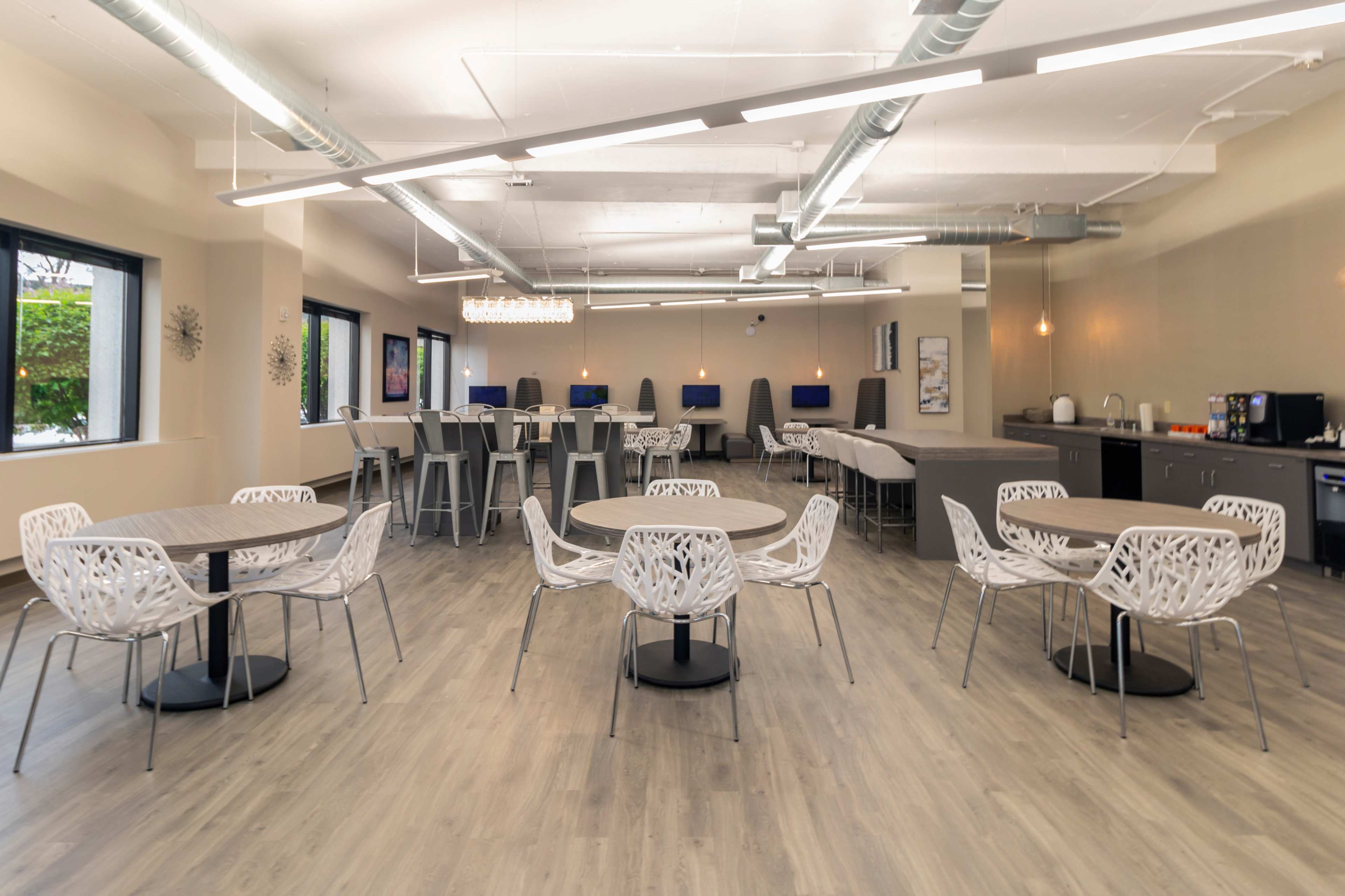 A spacious, modern cafeteria with several round tables and white chairs, featuring a kitchenette area and large windows.
