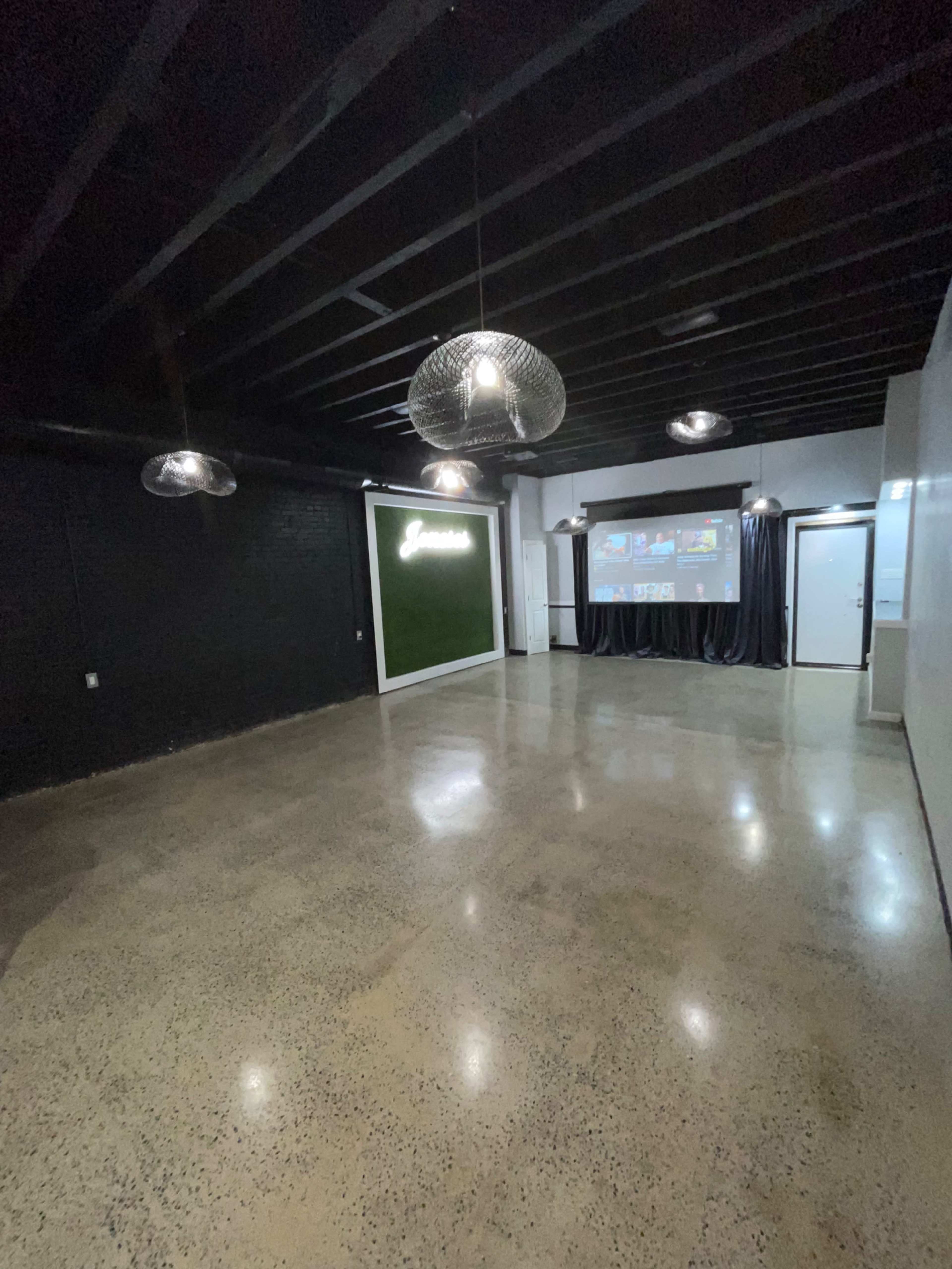 An empty room with polished concrete flooring, black walls, and industrial-style light fixtures hanging from the ceiling, featuring a projector screen on one wall.