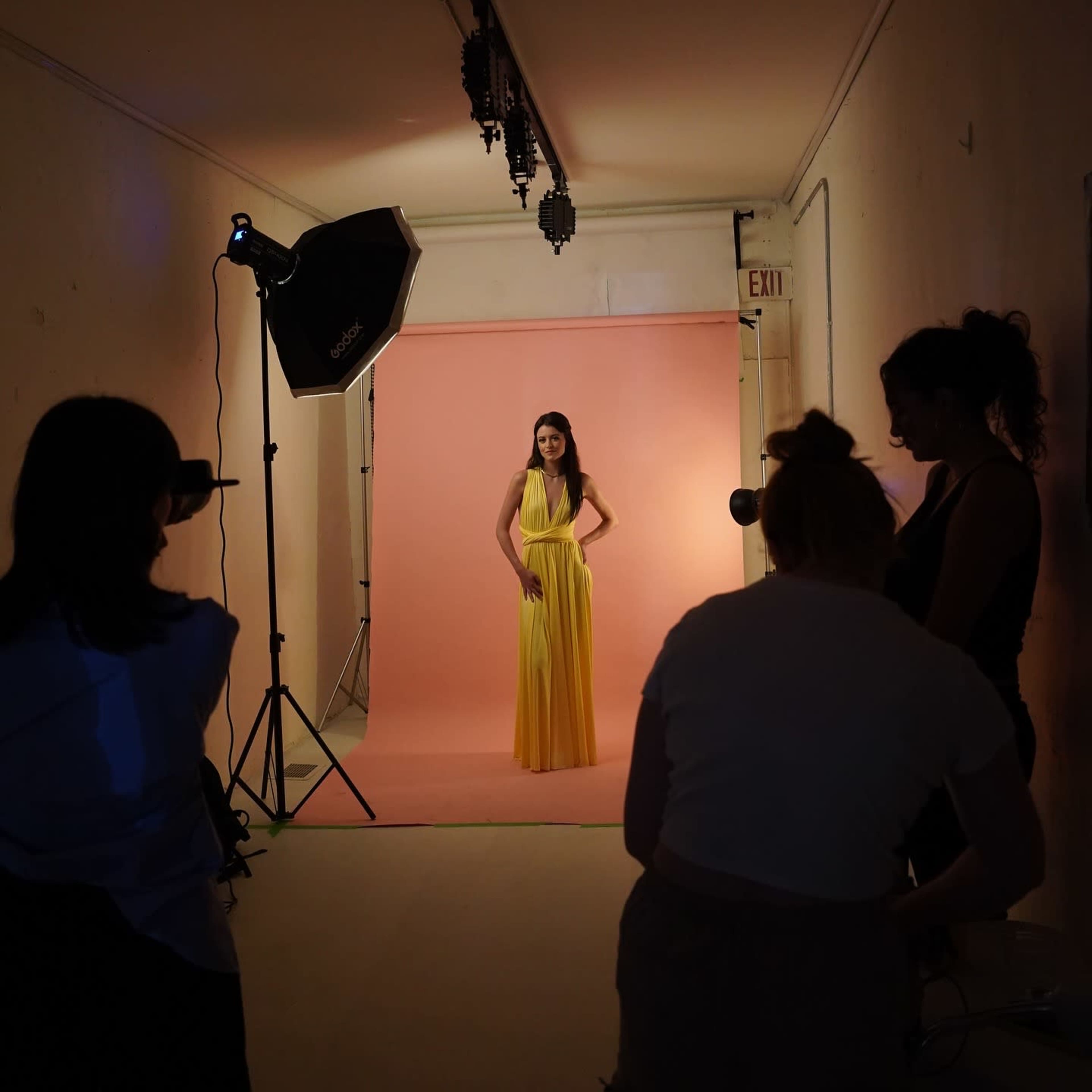 A model in a yellow dress poses in front of a pink backdrop while a small team adjusts lighting and photography equipment in a studio.