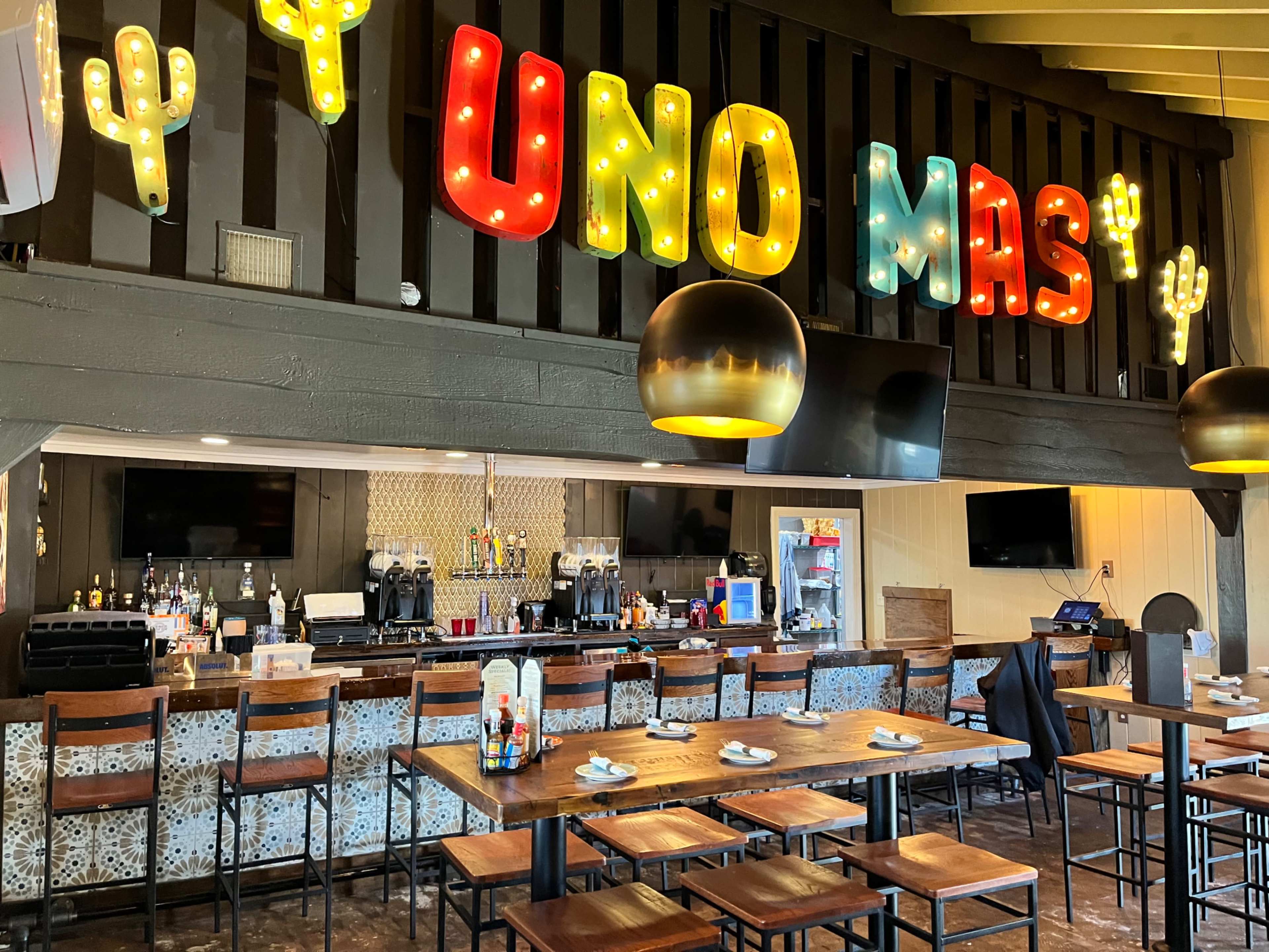 A brightly lit restaurant bar area with a sign that says "UNO MAS," wooden tables, and a variety of seating options.