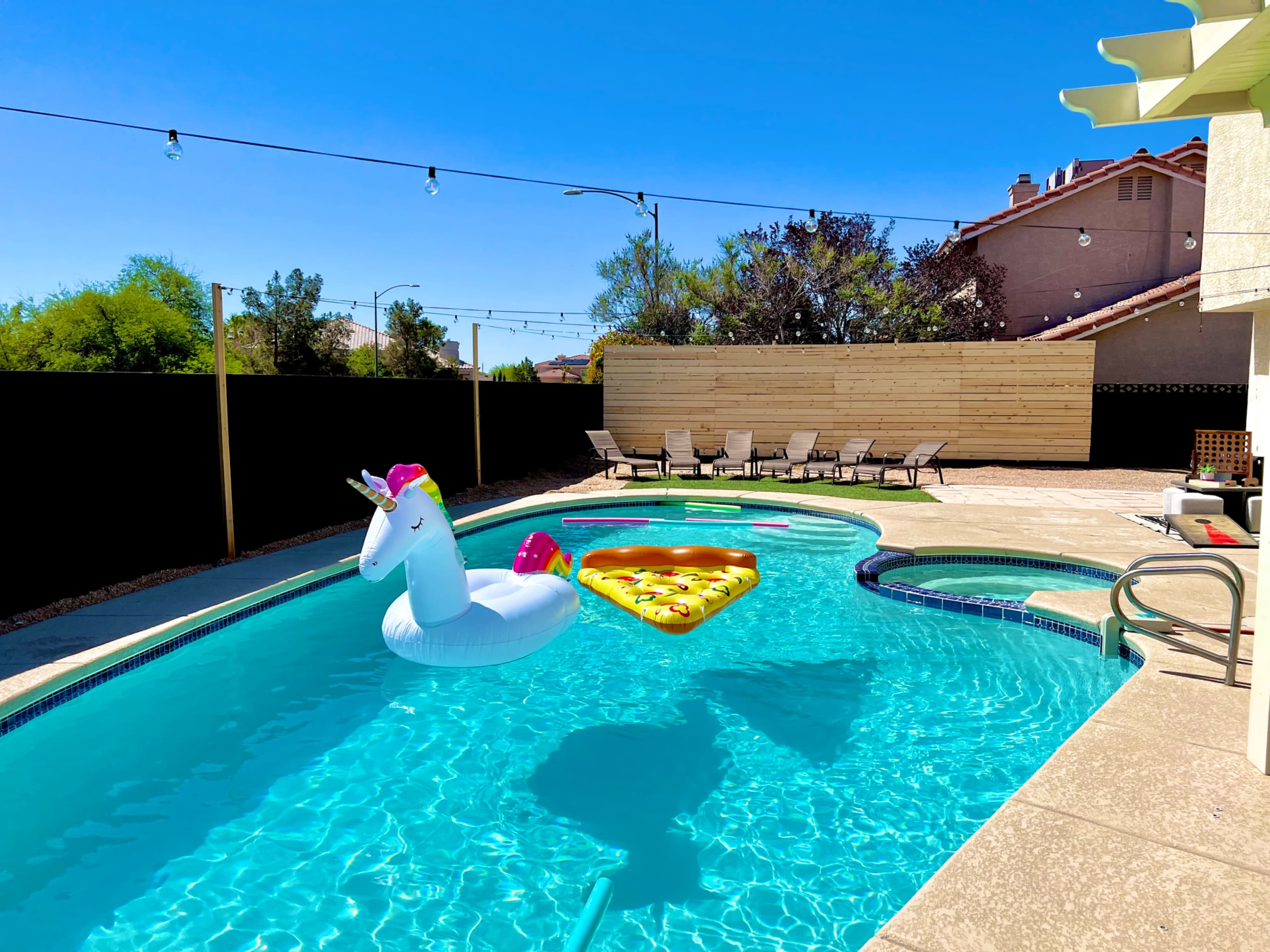 A unicorn float and a pizza-shaped float rest in a residential swimming pool surrounded by lounge chairs and a fenced yard under a clear blue sky.