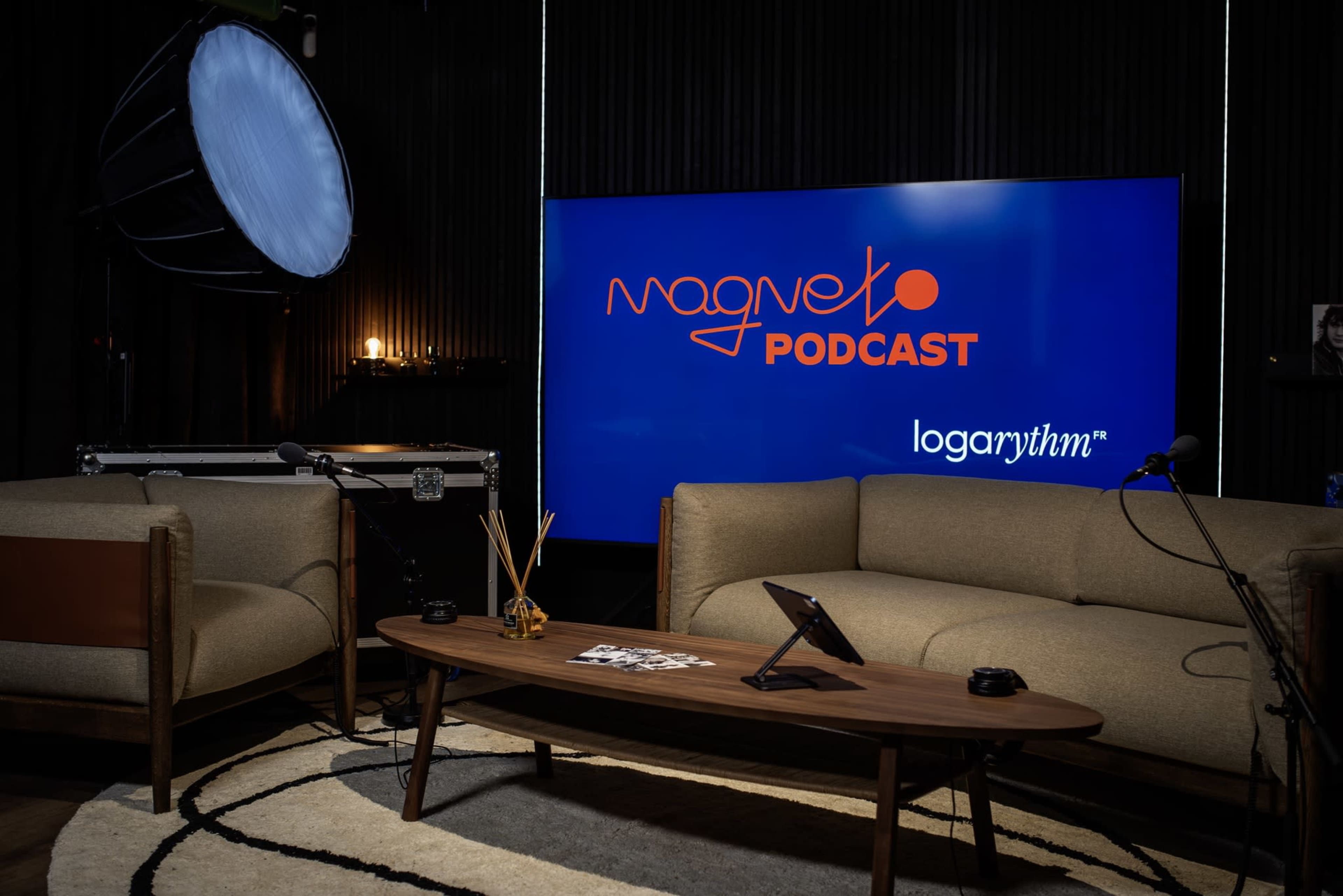 The image features a podcast recording set with two couches, a coffee table, and a large screen displaying the logo "Magneto Podcast."