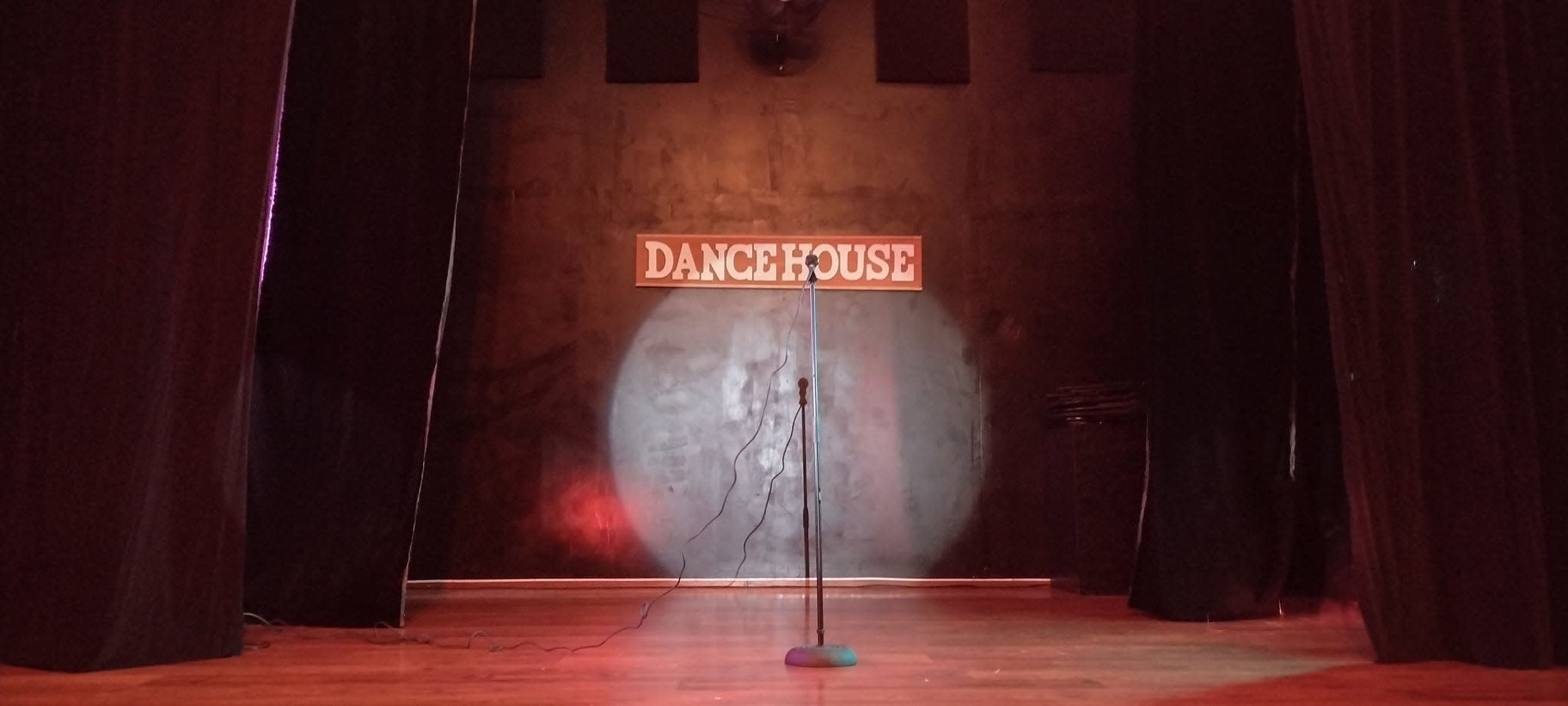 A microphone stands alone on a wooden stage under a spotlight, with a sign reading "DANCE HOUSE" on the back wall.