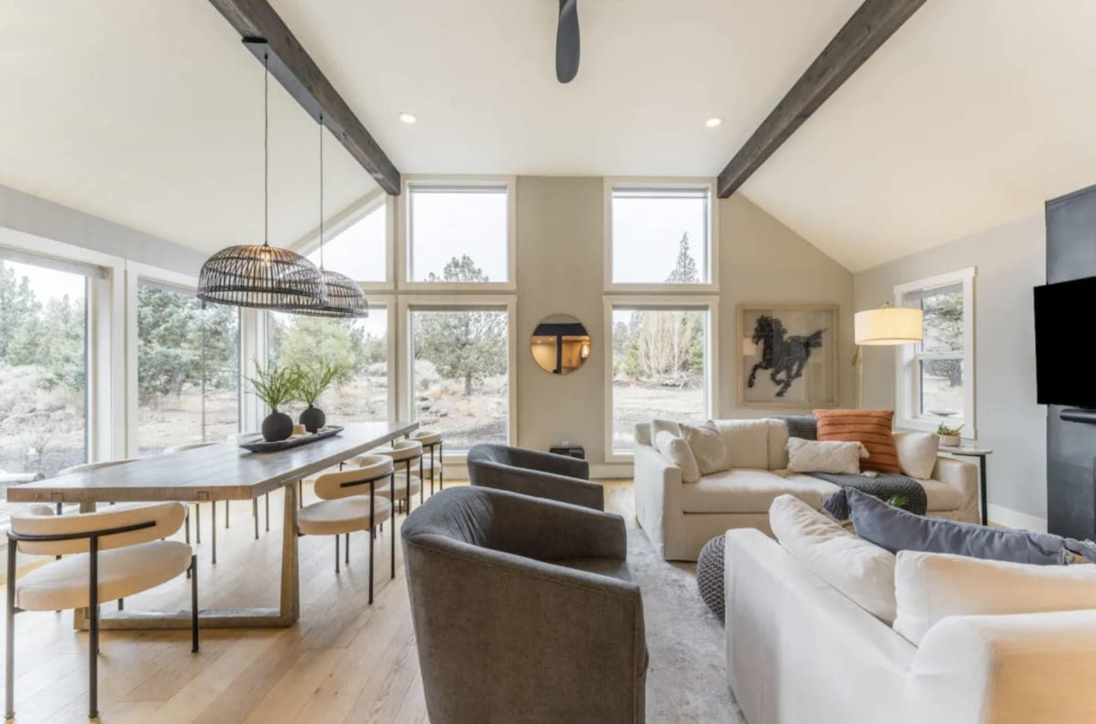 A modern living space features large windows, a wooden dining table, comfortable seating, and a flat-screen TV.