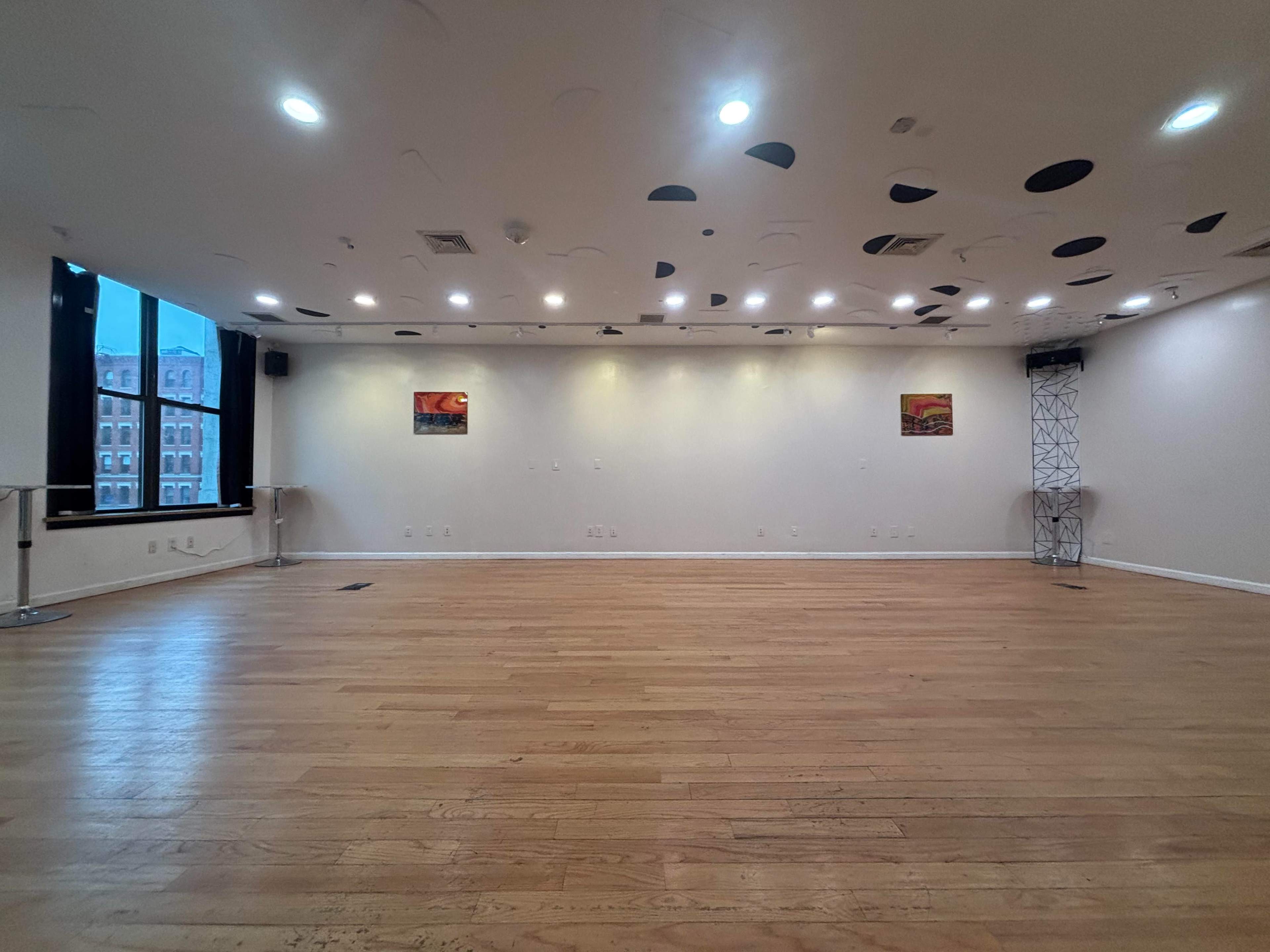 The image shows a spacious, well-lit dance studio with a wooden floor, large windows, and paintings on the walls.