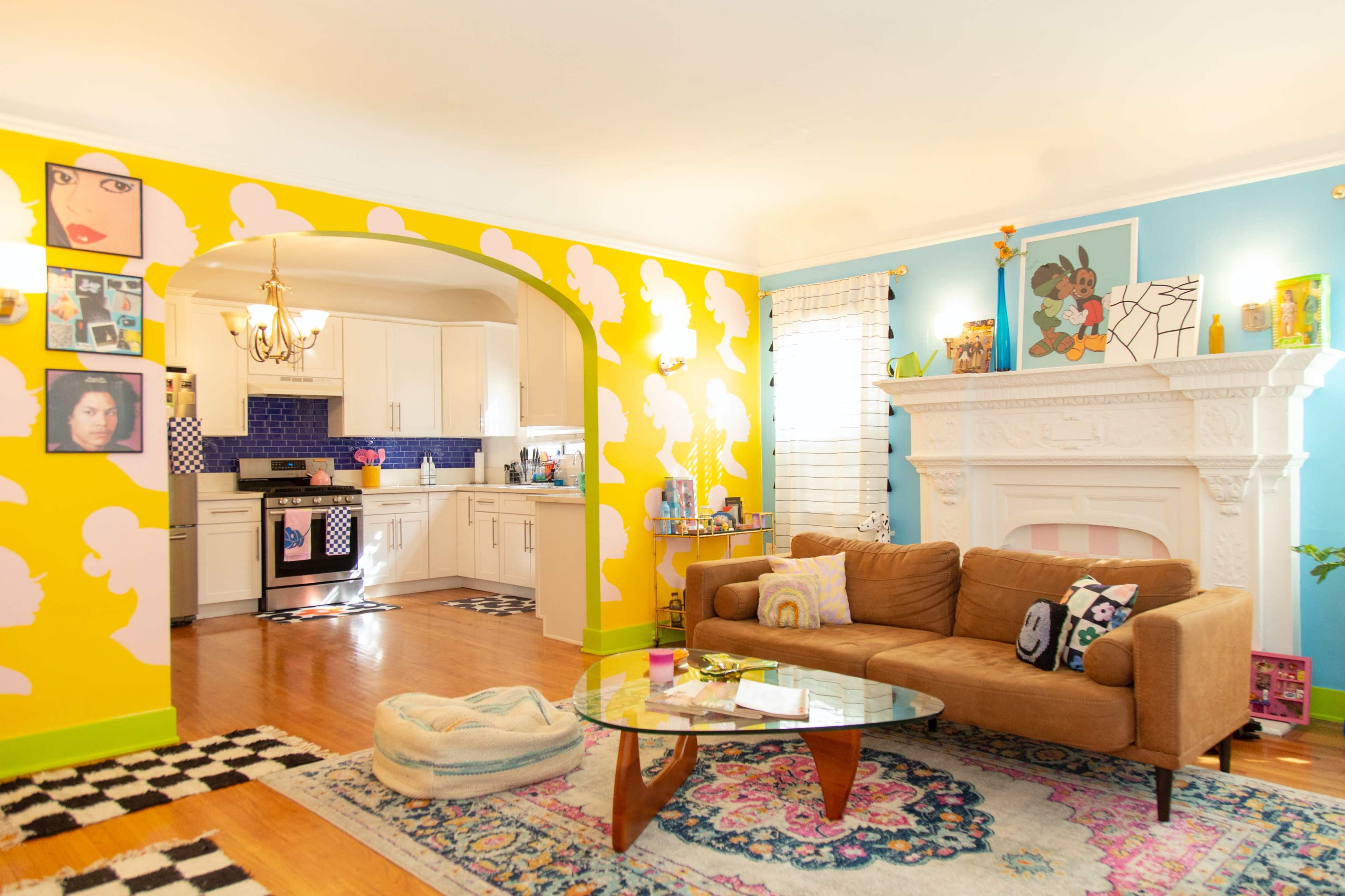The living room features a brightly painted yellow wall with cloud patterns, a cozy brown sofa, a glass coffee table, and a white fireplace surrounded by colorful artwork.