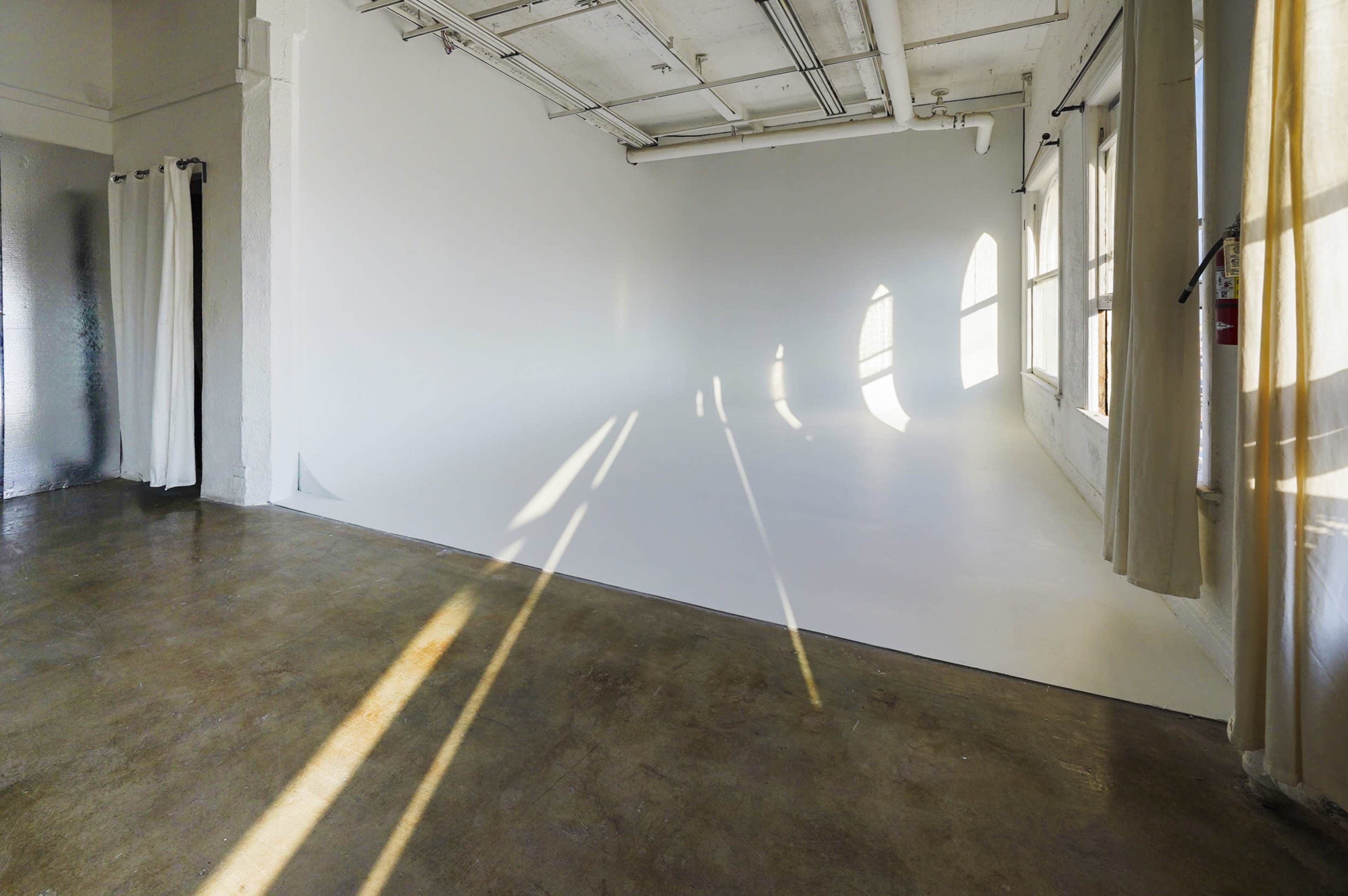 The image shows a minimalist studio space with white walls, a light-colored floor, and sunlight casting shadows from the windows.