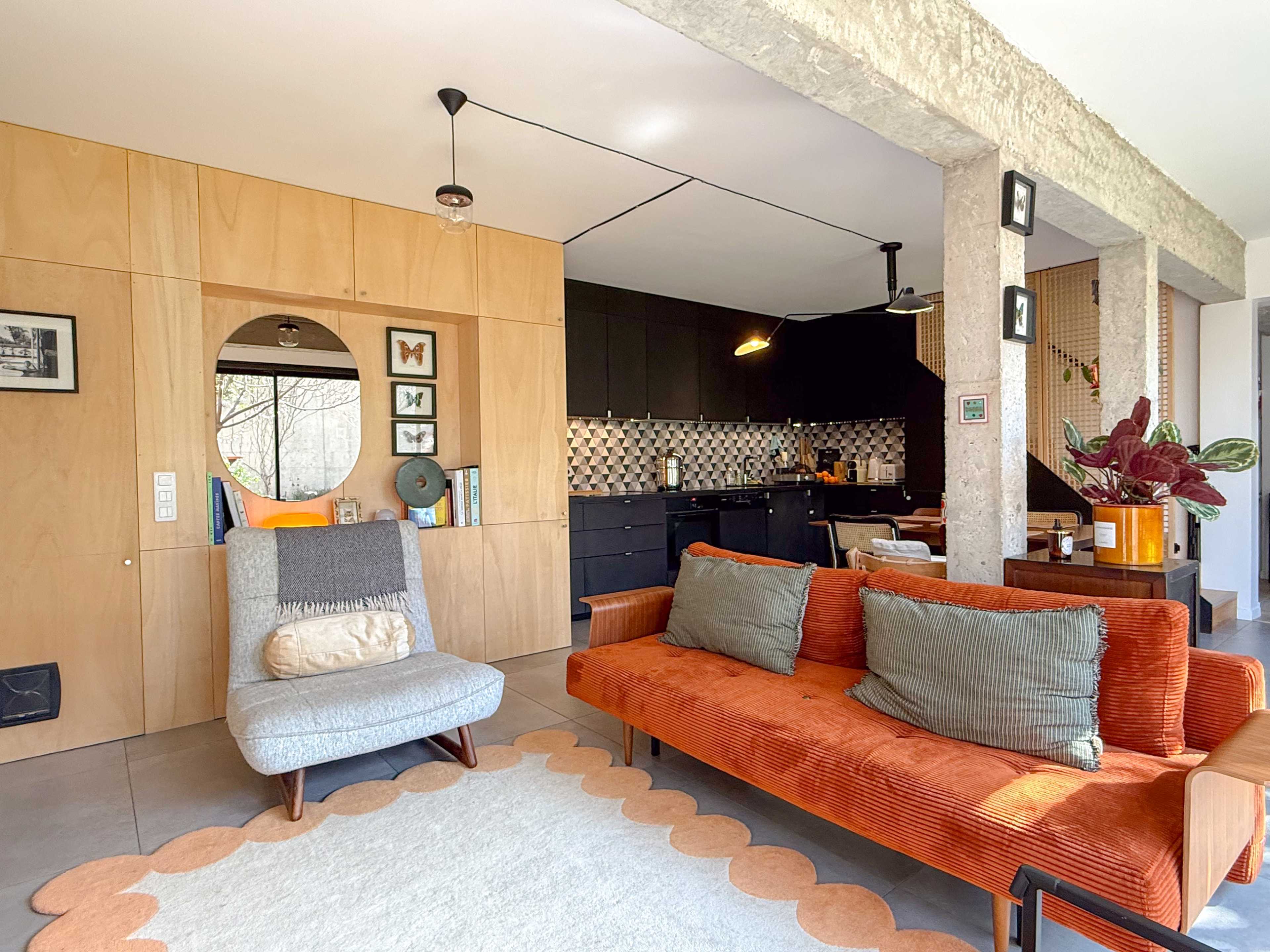 The image shows a contemporary living space with a mix of wooden and concrete elements, featuring a stylish orange sofa, a gray chair, and an open kitchen in the background.