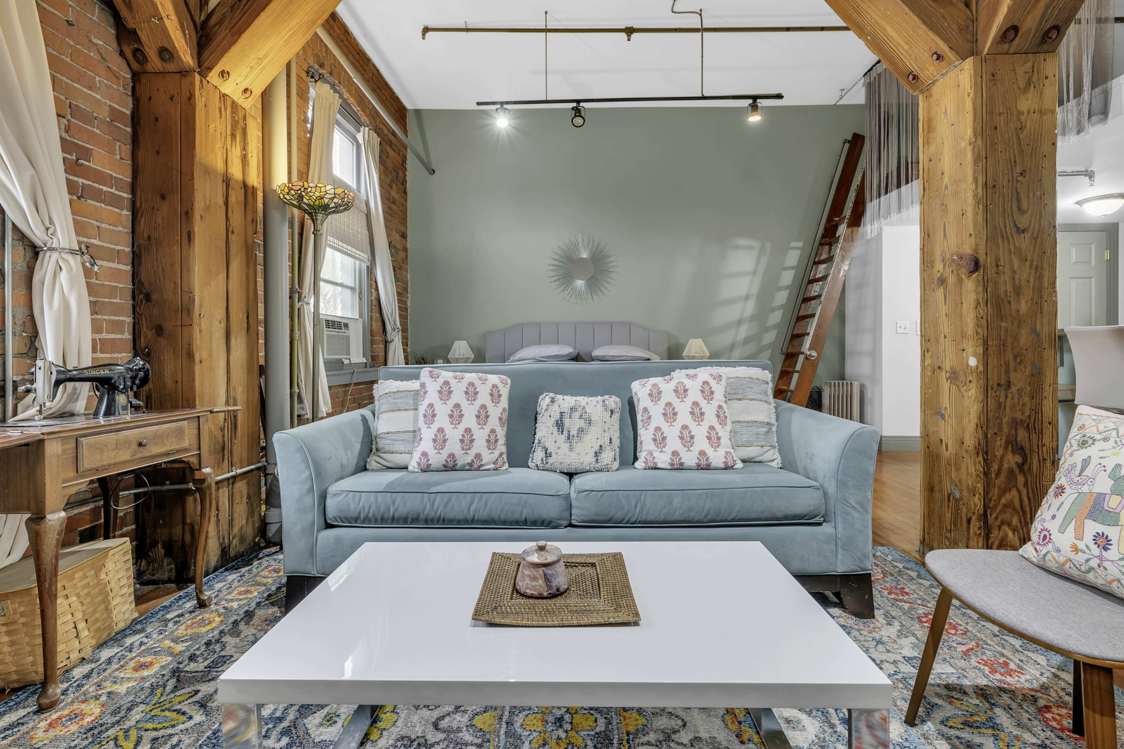 A cozy living area features a light blue sofa with decorative pillows, a white coffee table, exposed wooden beams, and a ladder leading to a bed in the background.