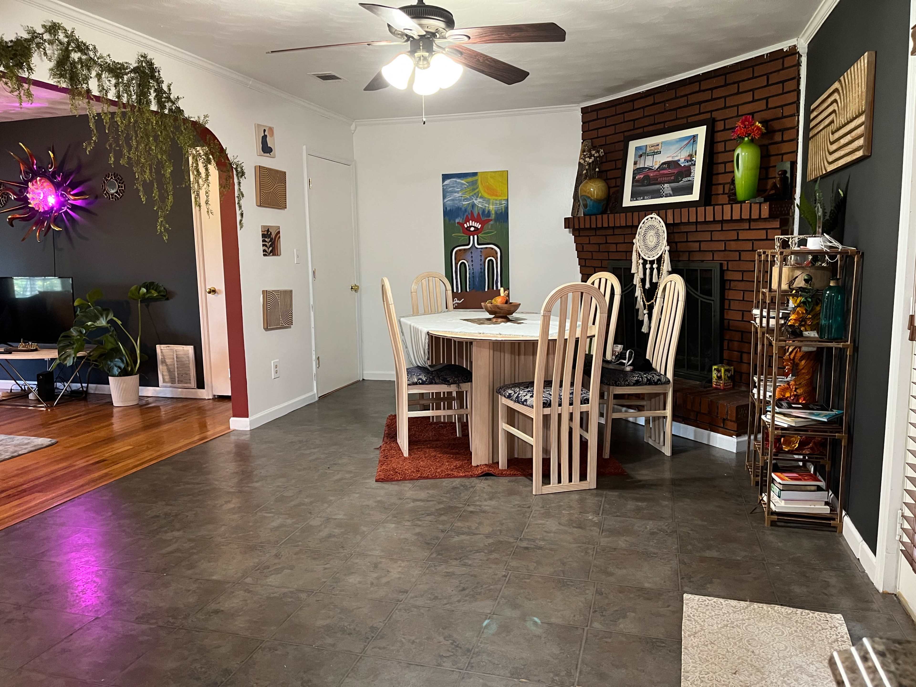 The image shows a spacious dining area with a round table surrounded by six chairs, a brick fireplace adorned with decorations, and a colorful artwork on the wall.