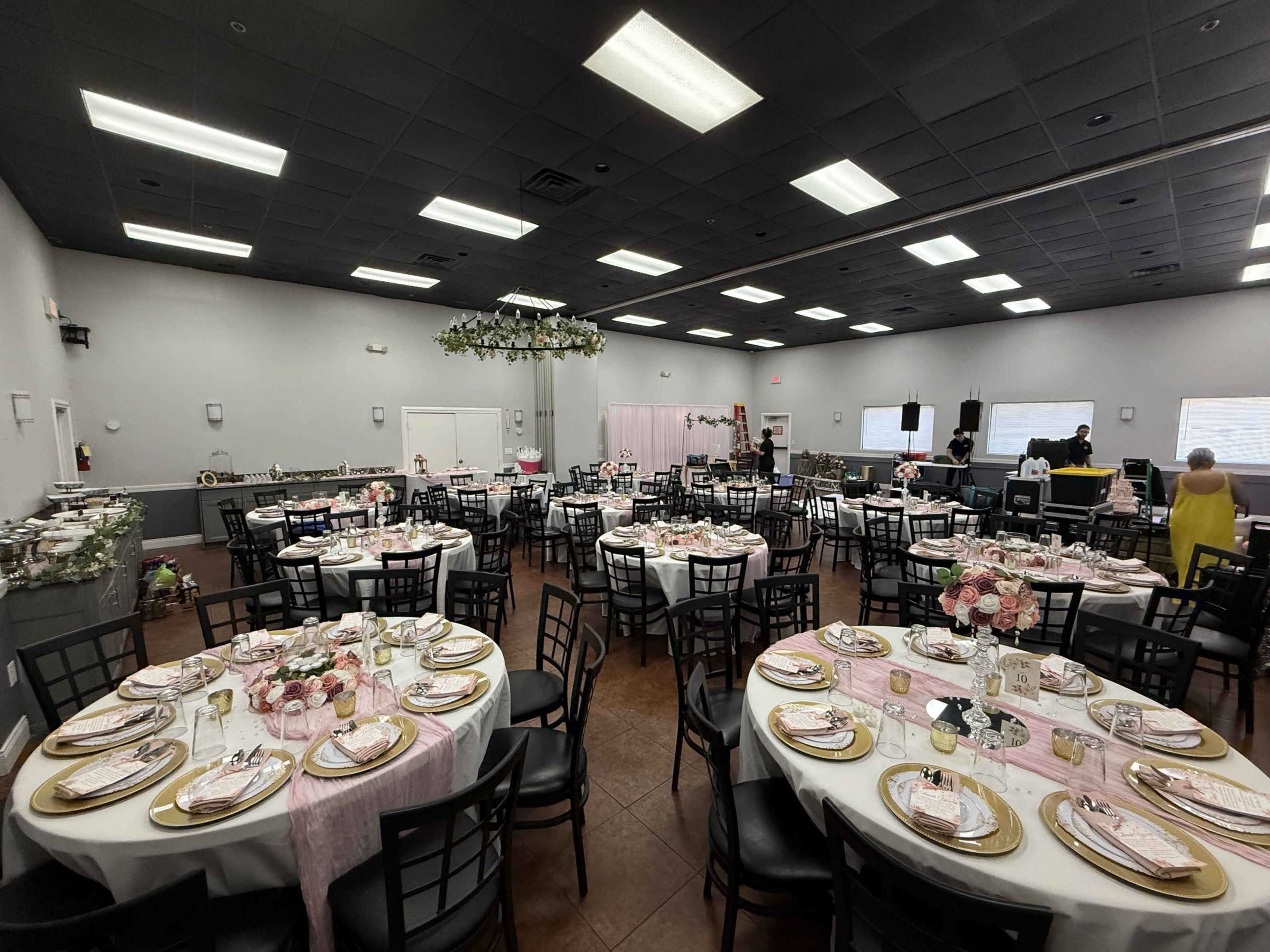 The image shows a banquet hall arranged for an event, featuring round tables set with pink and gold tableware, floral centerpieces, and a decorative ceiling arrangement.