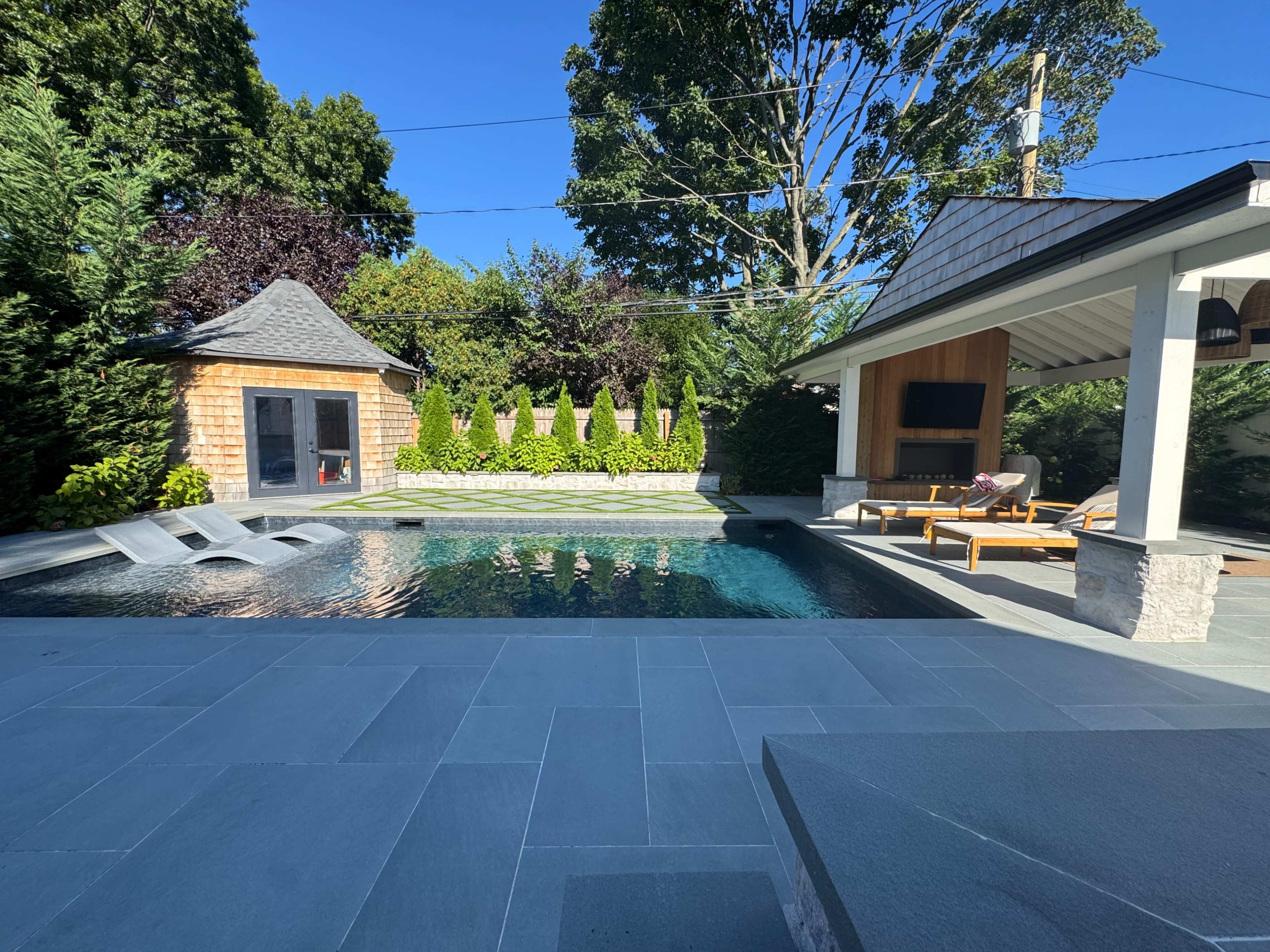 The image displays a backyard pool area featuring a modern swimming pool, lounge chairs, a covered structure with an outdoor fireplace, and landscaped greenery surrounding the space.