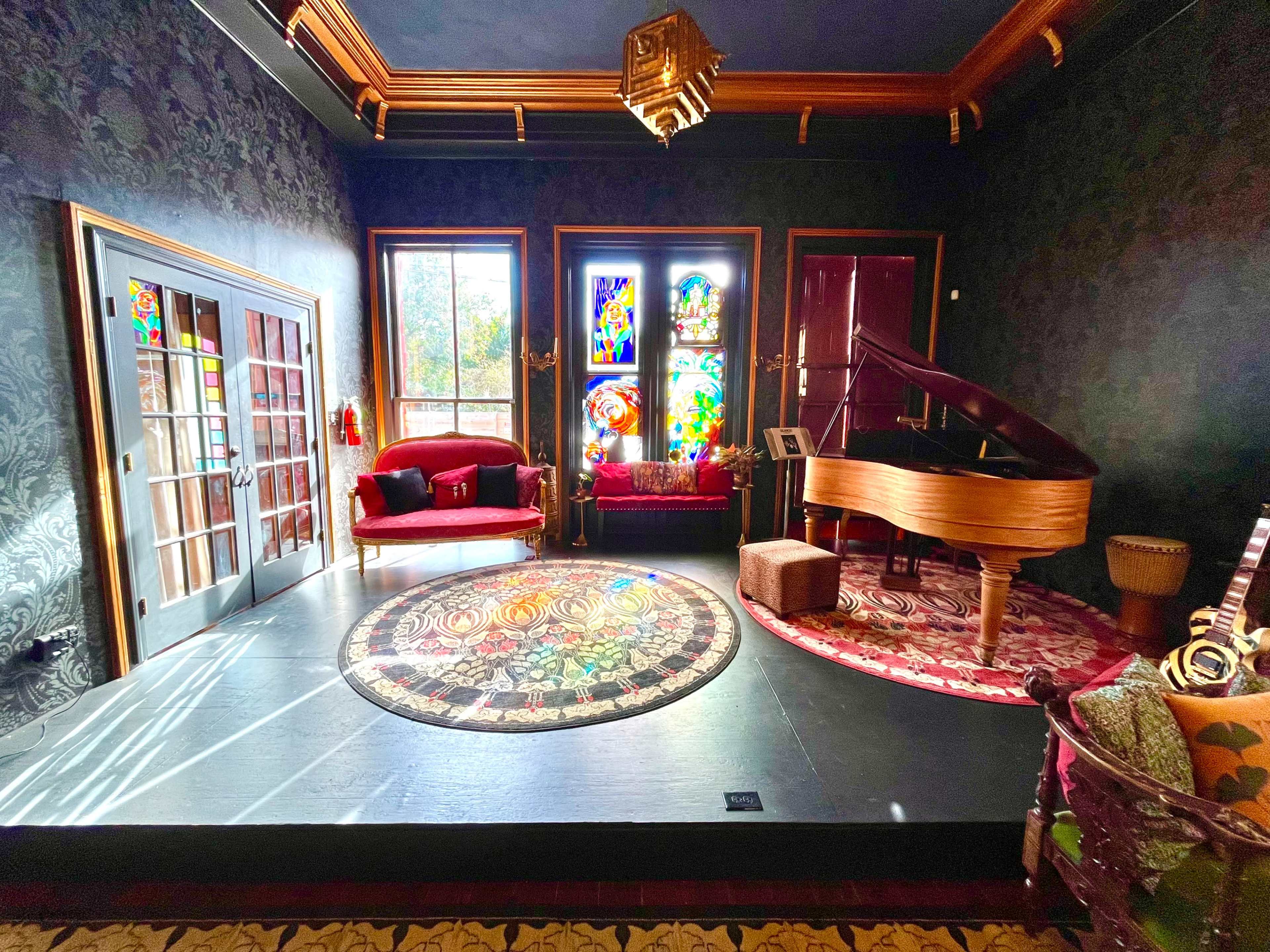 A well-furnished room featuring a grand piano, a red velvet sofa, and large stained glass windows, with intricate rugs on the floor.