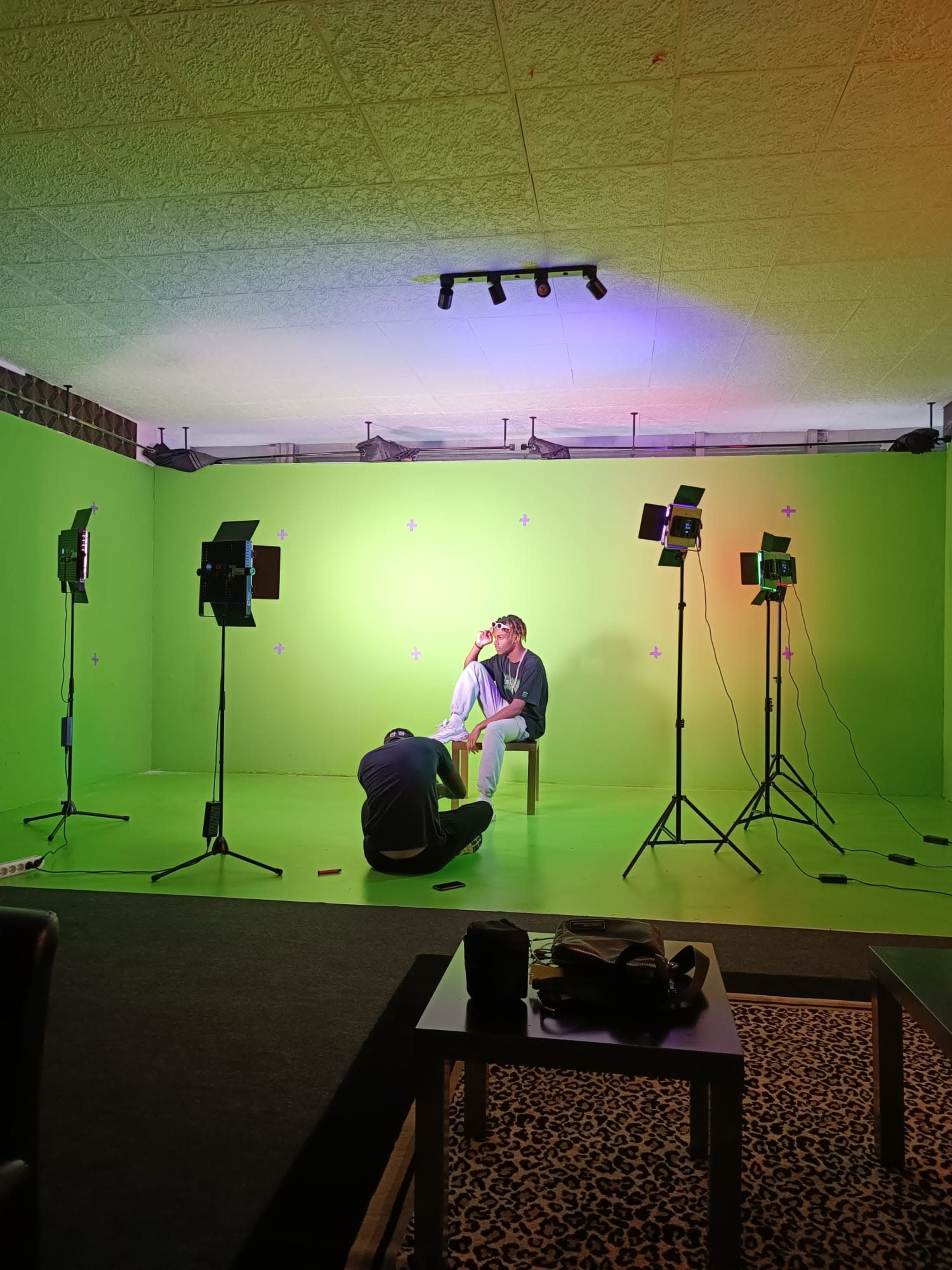 A person sits on a chair in a brightly colored green room surrounded by several studio lights and another individual on the floor nearby.