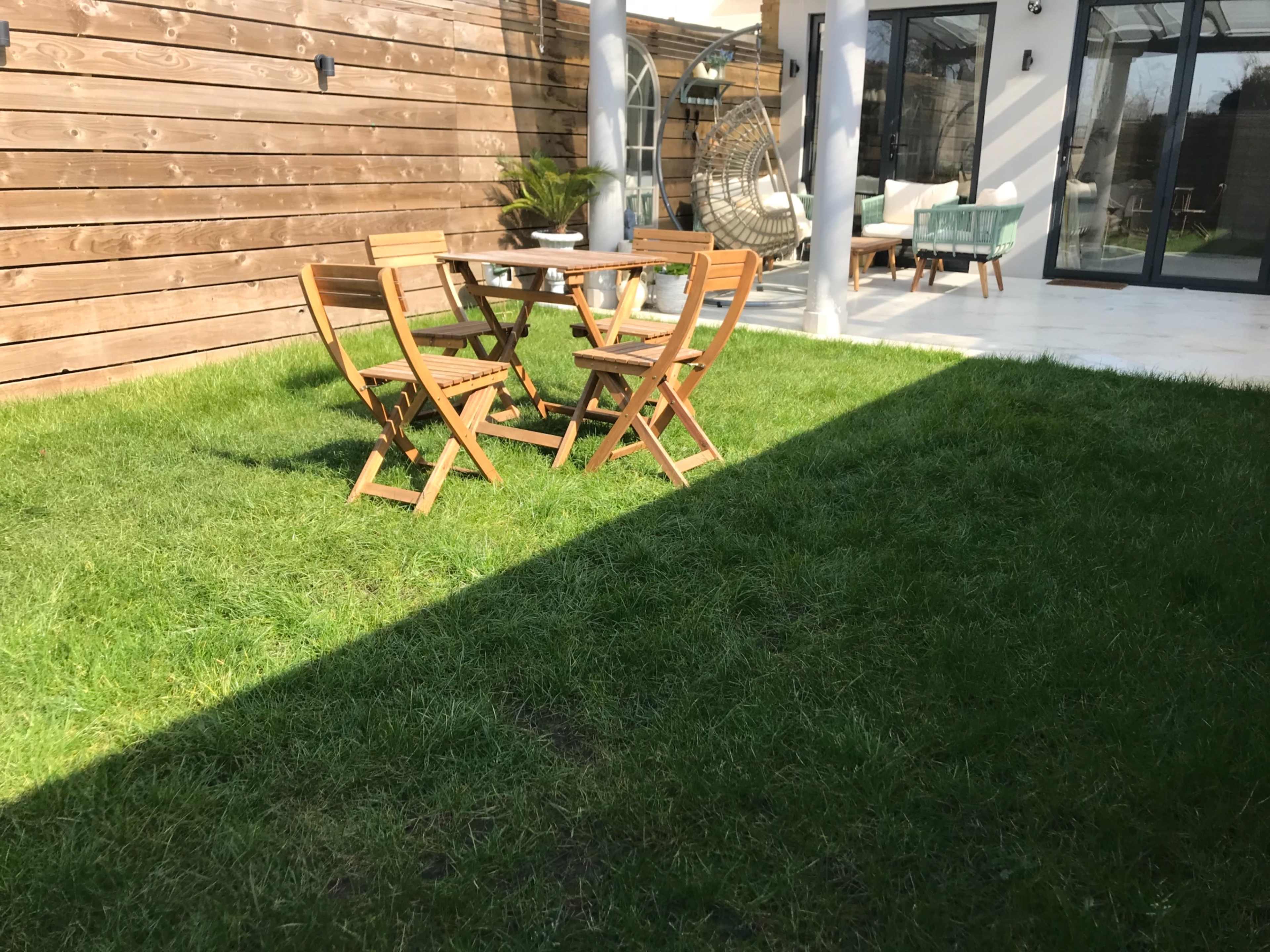 A small outdoor seating area with four wooden chairs and a round table is situated on a green lawn beside a modern glass-walled building.