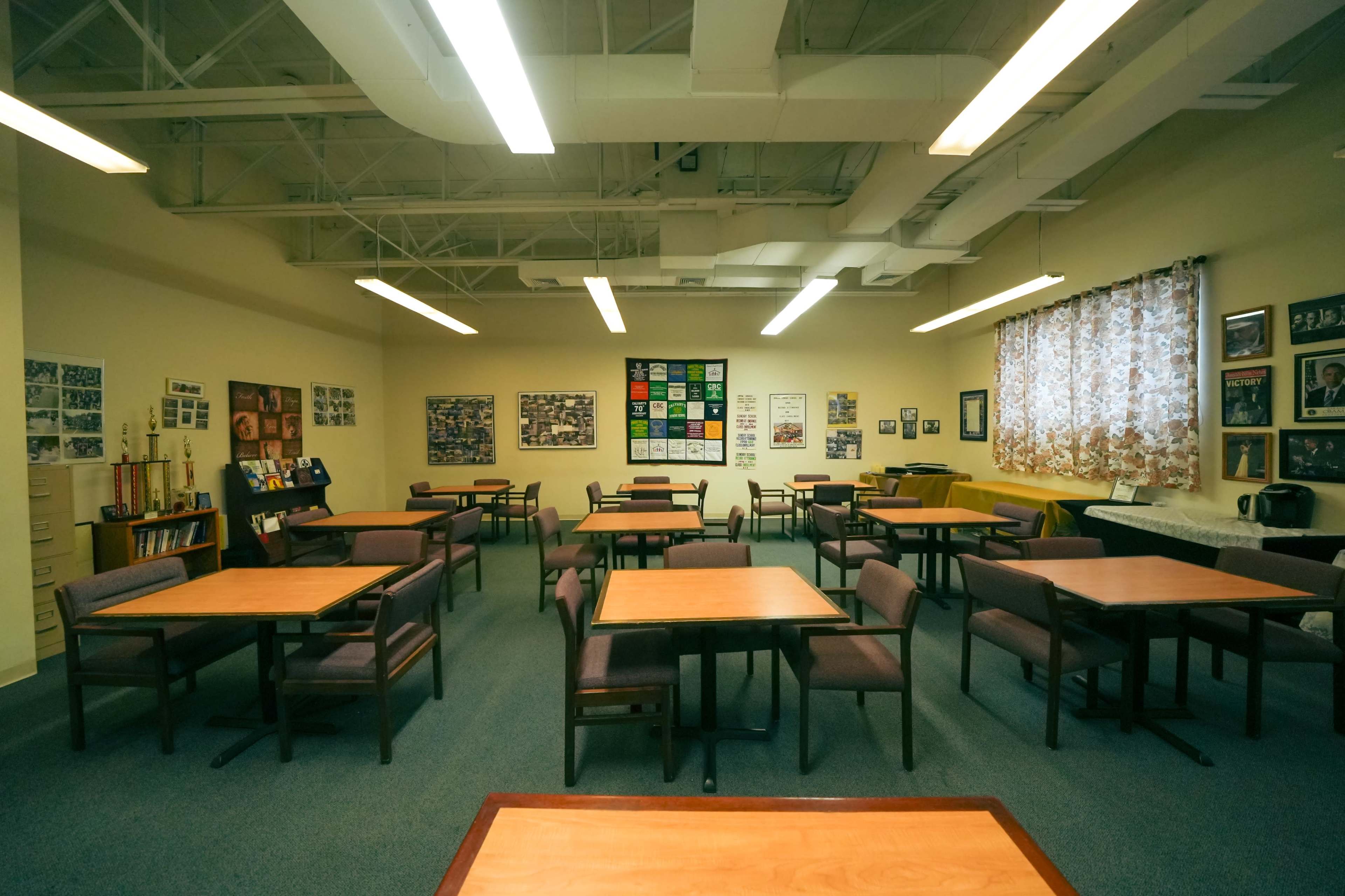 A classroom with several empty tables and chairs arranged in a spacious layout, surrounded by walls adorned with photographs and posters.