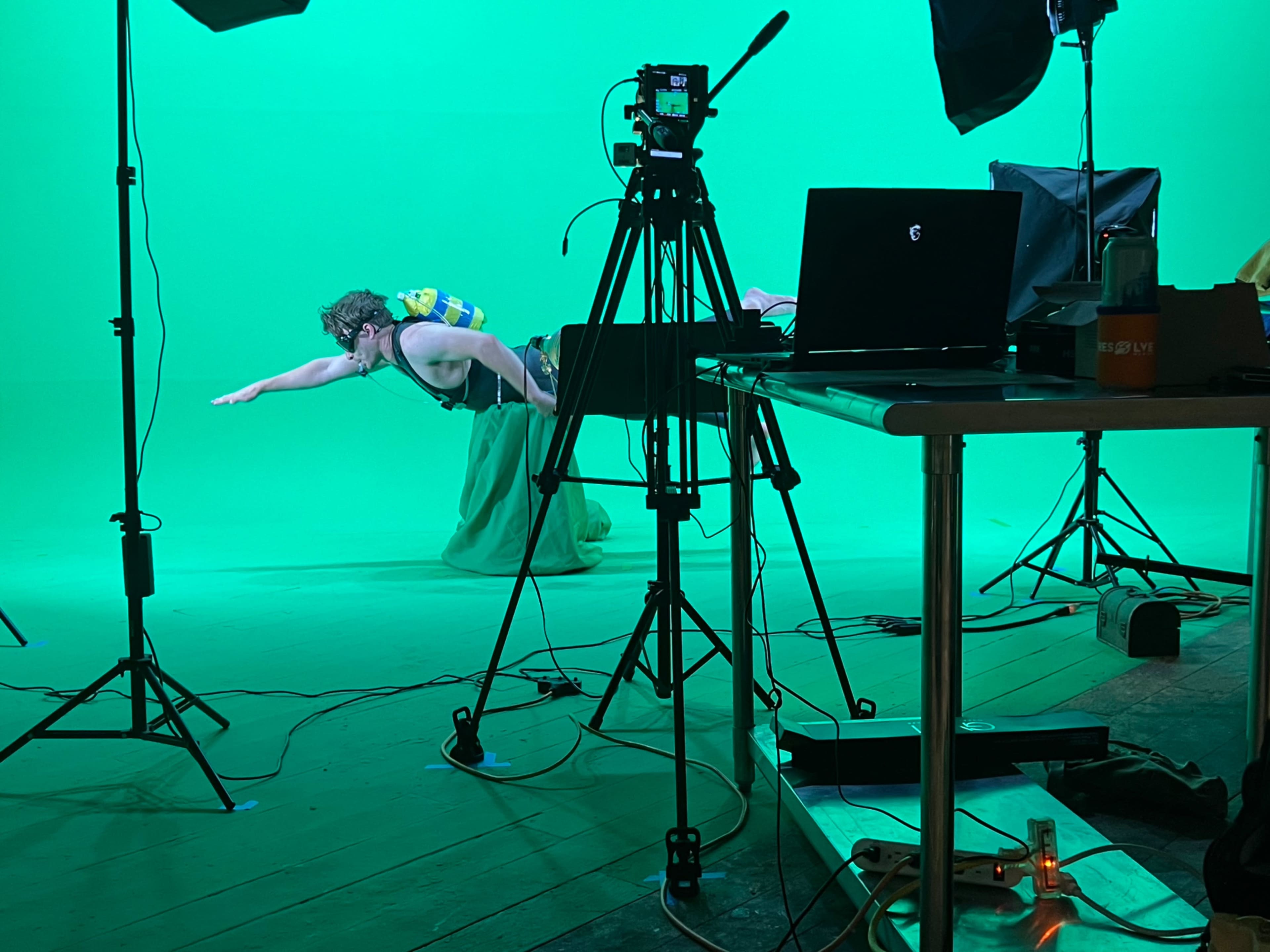A person is swimming in an indoor studio with a green screen backdrop, surrounded by camera equipment and a laptop on a table.