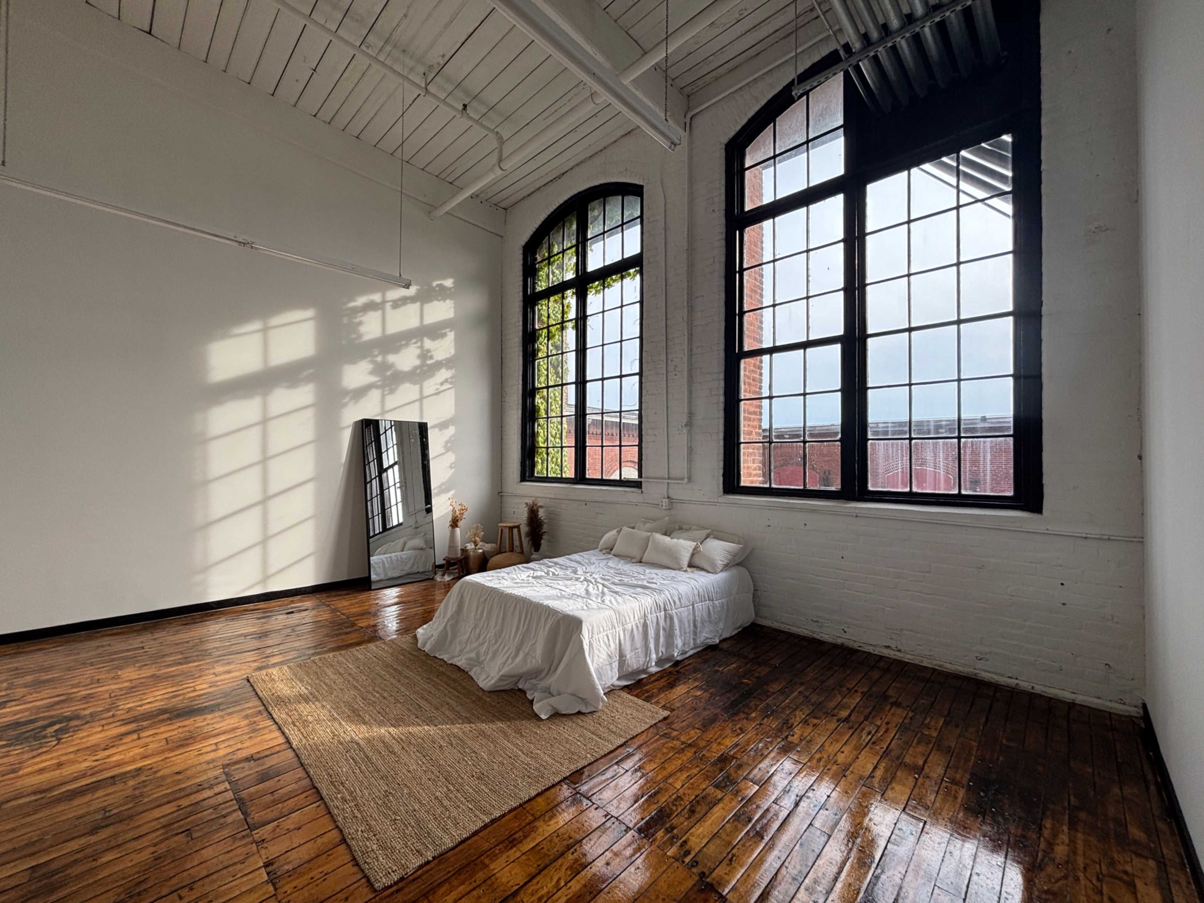 A minimalist bedroom features a bed with white linens, a large mirror, and tall windows that cast shadows on the wooden floor.