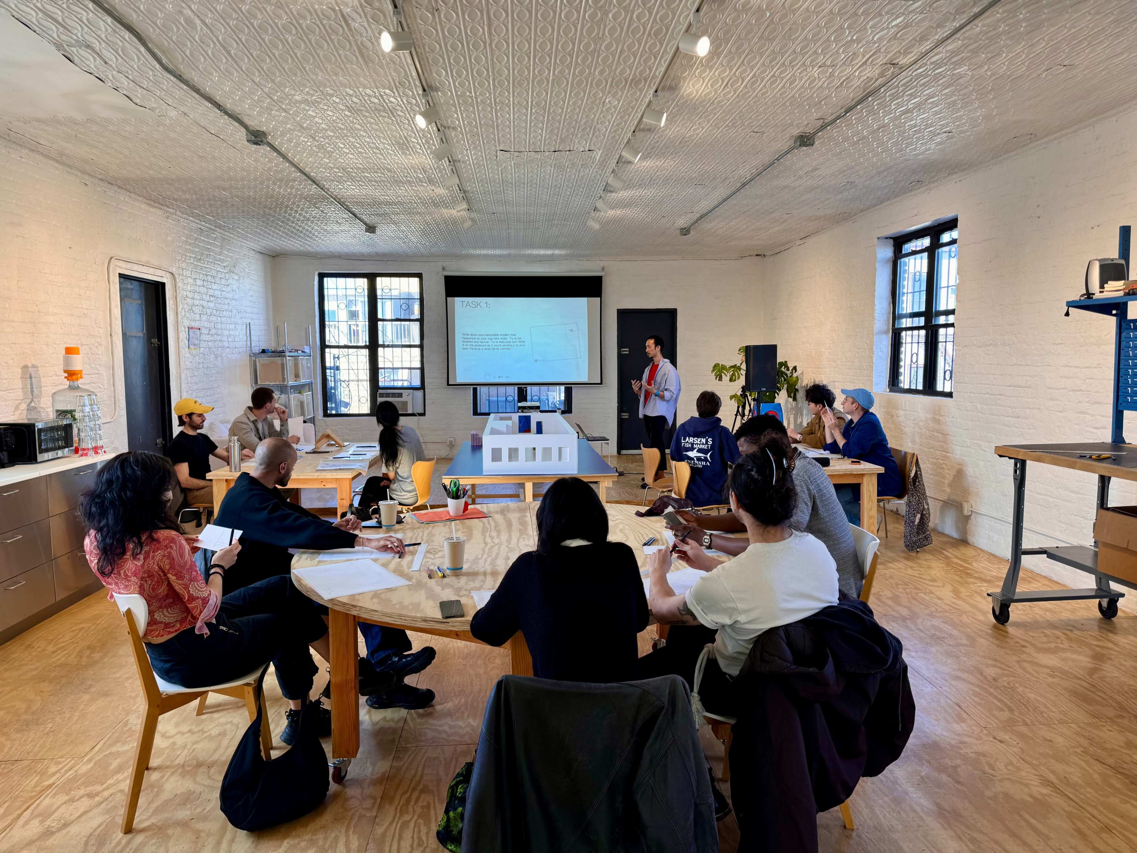 A group of people sits around tables in a bright room while a presenter stands in front of a screen.