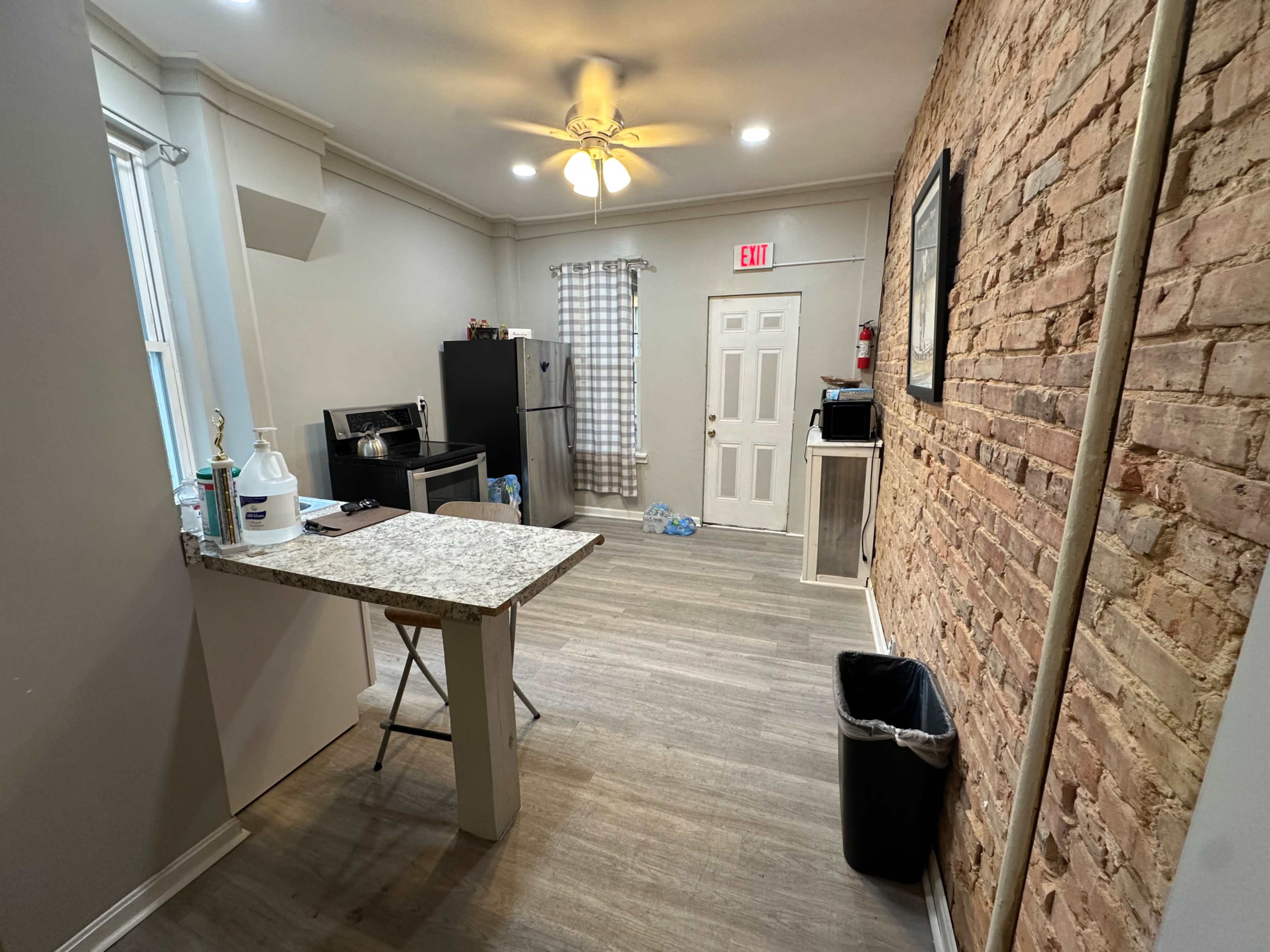 The image shows a kitchen area featuring a countertop with a few items, a black refrigerator, and a door leading to another room, with exposed brick walls and light-colored flooring.