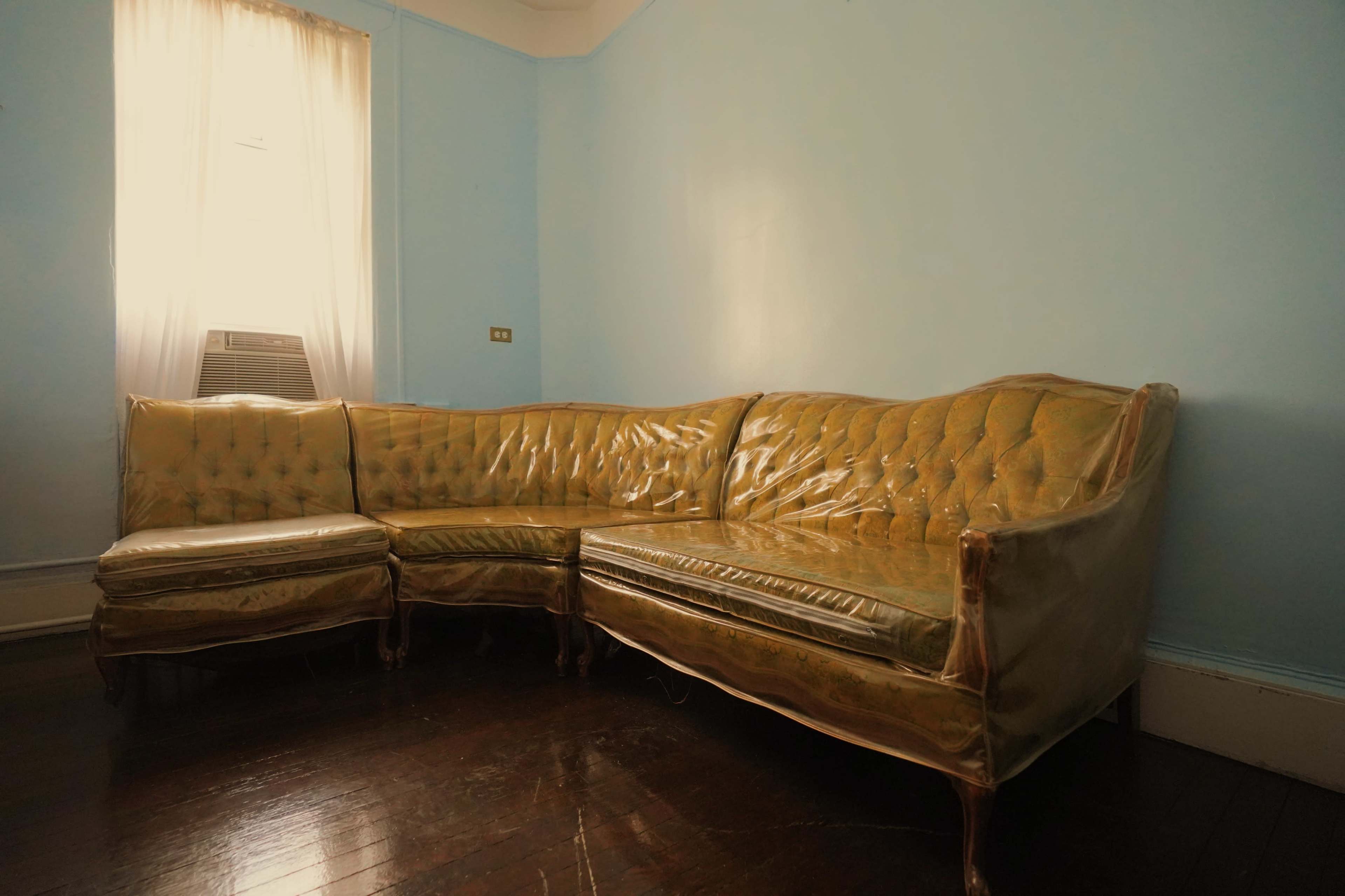 A vintage sectional couch is covered in plastic in a sparsely furnished room with light blue walls and a window.