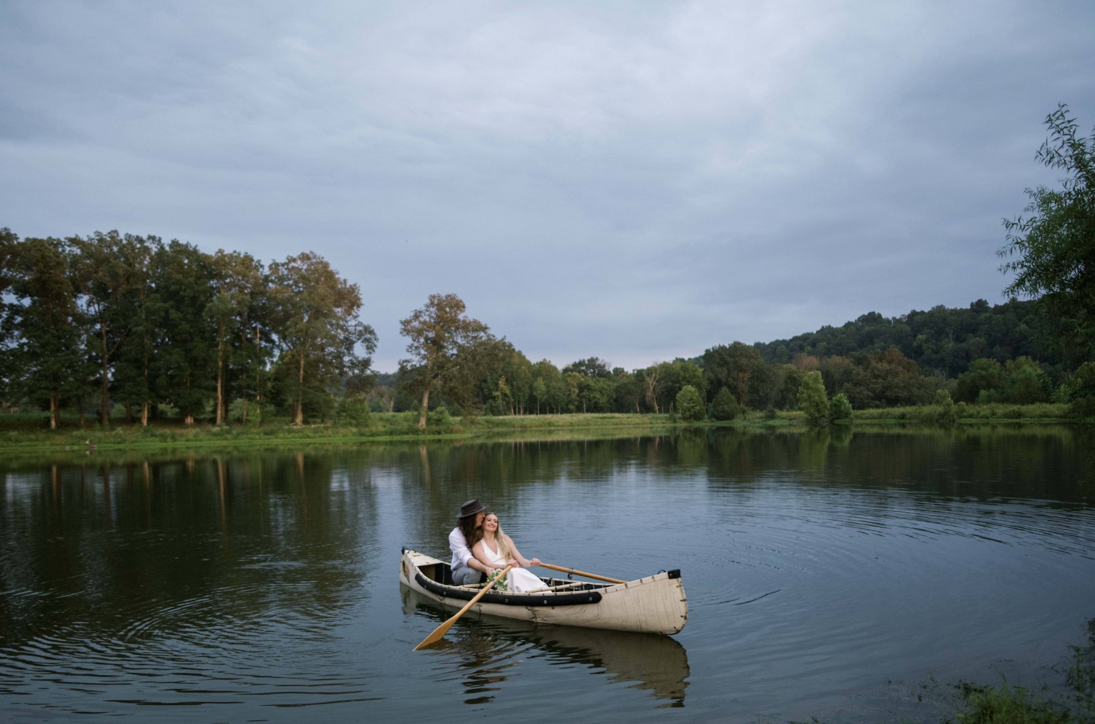 A couple rows a canoe across a calm lake surrounded by trees under a cloudy sky.