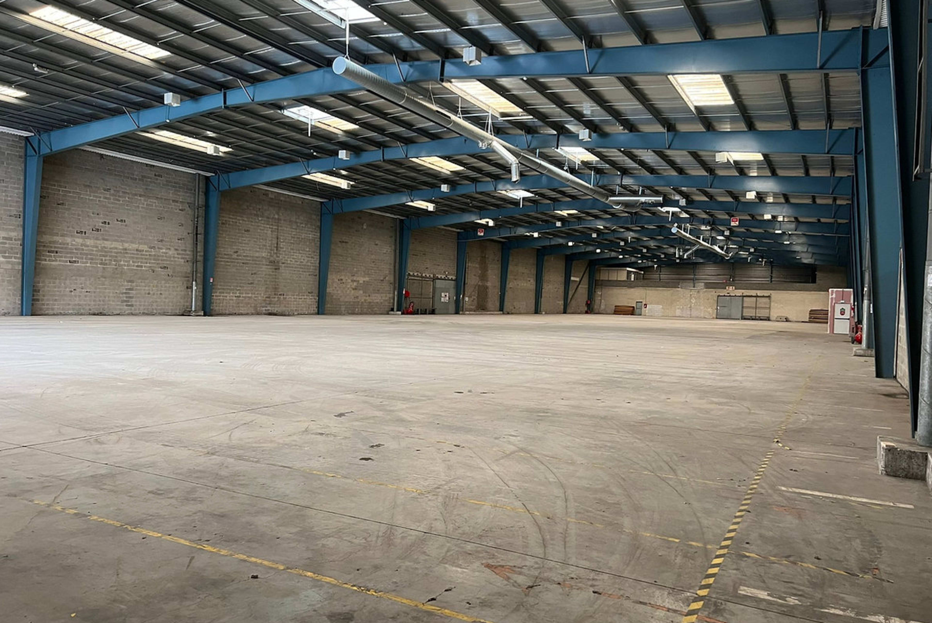 An empty industrial warehouse features high blue metal beams and a concrete floor with faint tire marks.