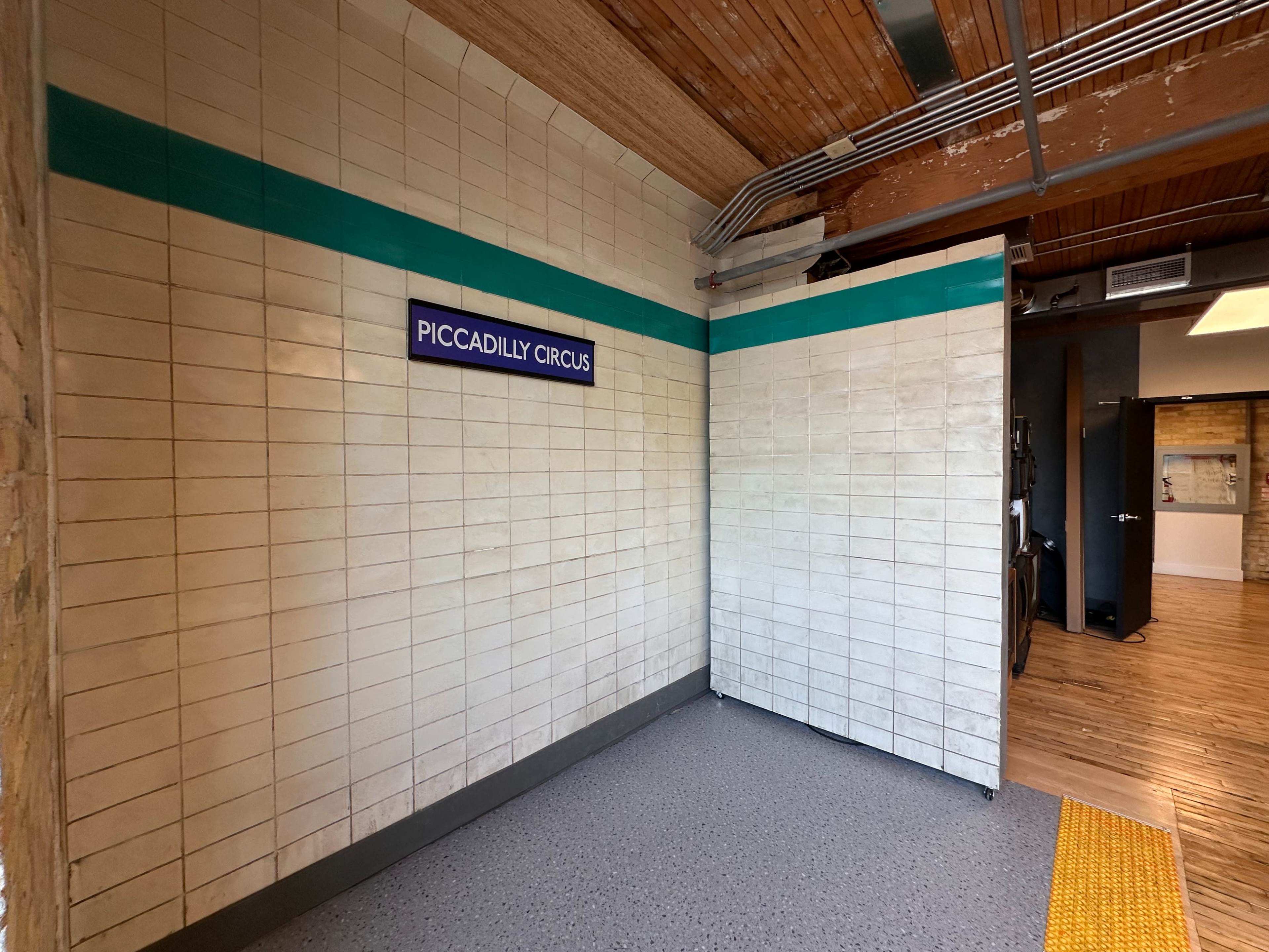 The image shows a tiled wall with a sign that reads "PICCADILLY CIRCUS" in a corner of a room, with wooden flooring visible beyond it.