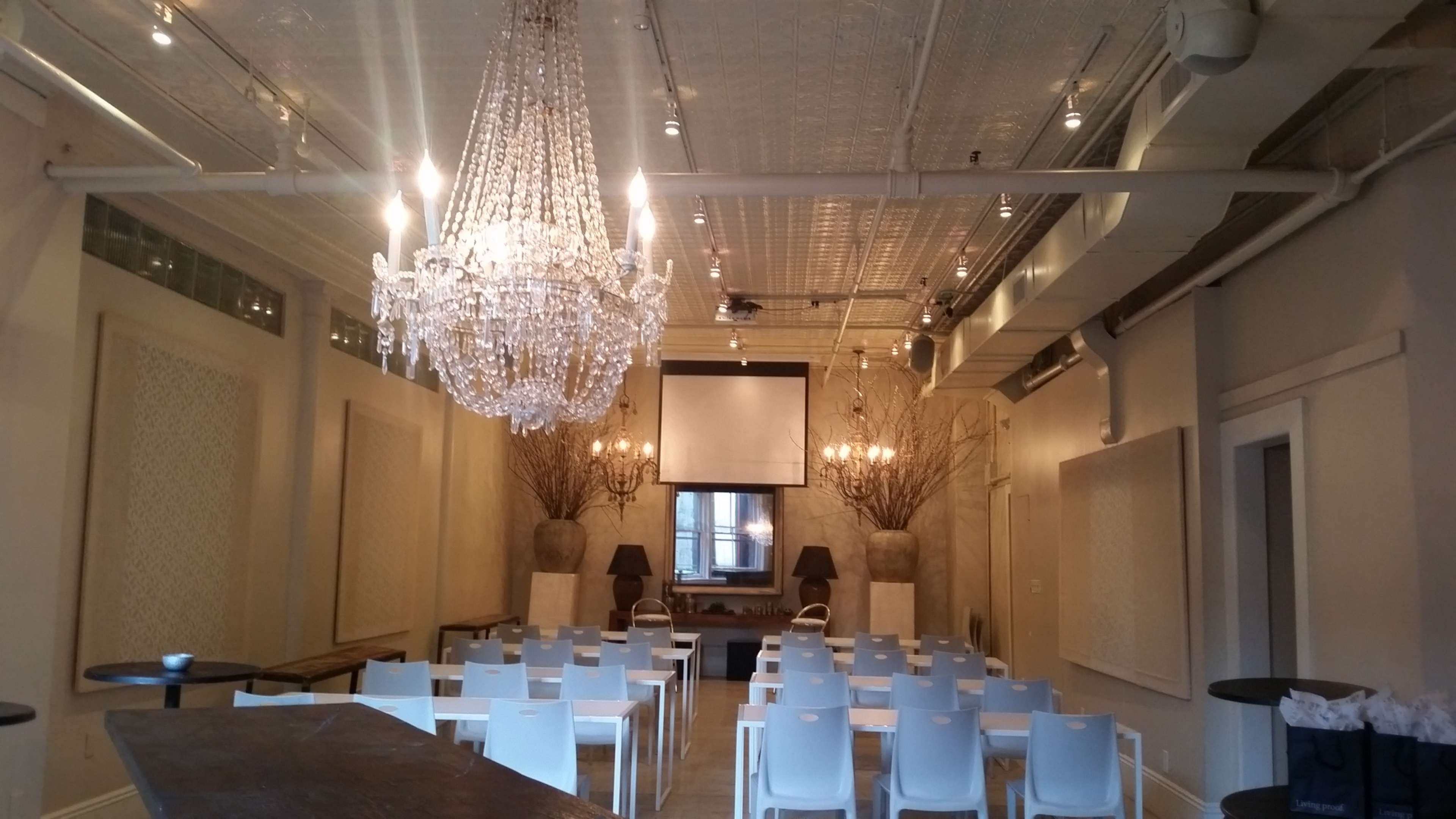 A spacious event room features a large chandelier, two decorative planters, and rows of white chairs arranged for a presentation.