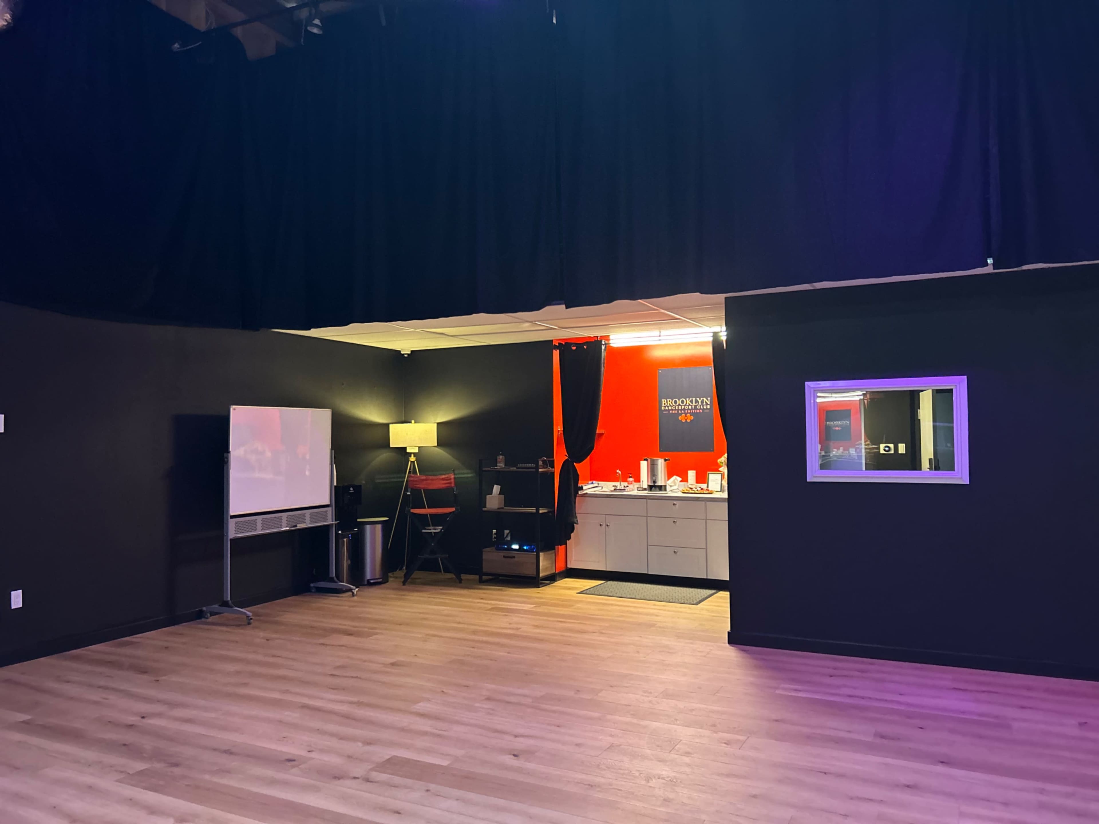 The image shows a spacious room with black walls, wooden flooring, and a kitchenette illuminated by red and white lighting.