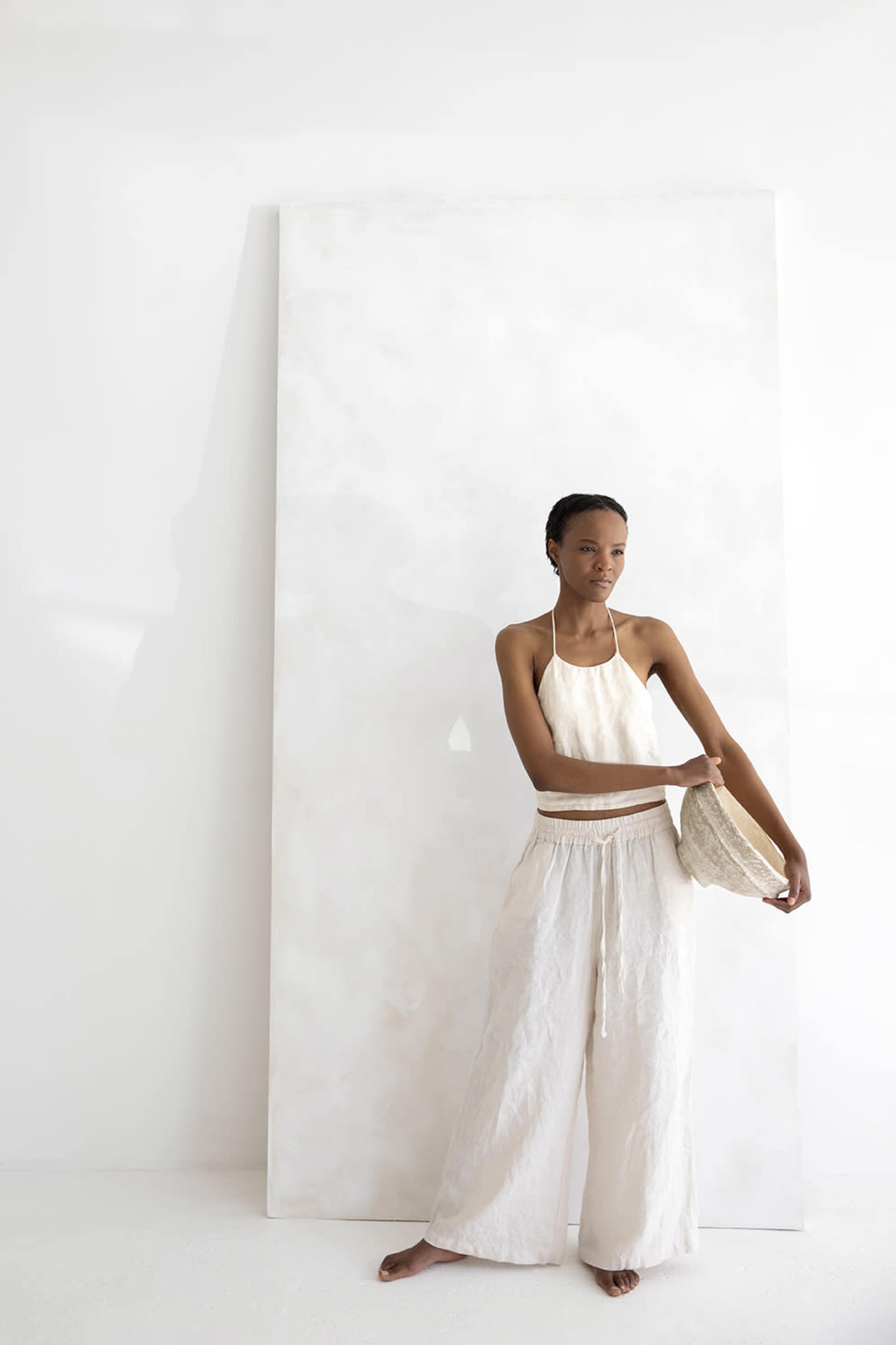 A woman stands barefoot next to a large white panel, wearing a light-colored halter top and wide-legged pants, while holding a woven bag.