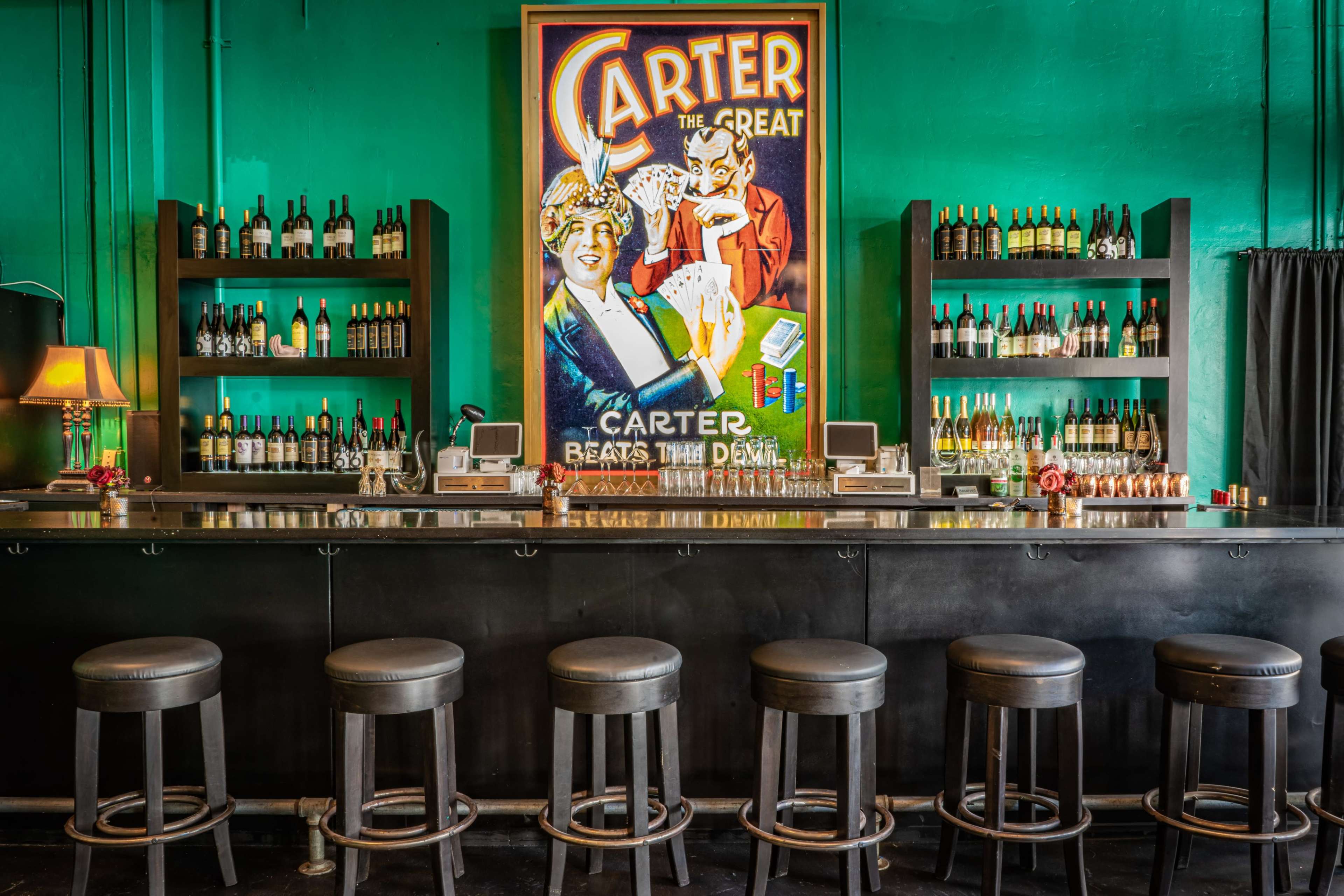 A bar with a green wall features a large vintage poster of a magician and shelves lined with various bottles of wine and liquor.