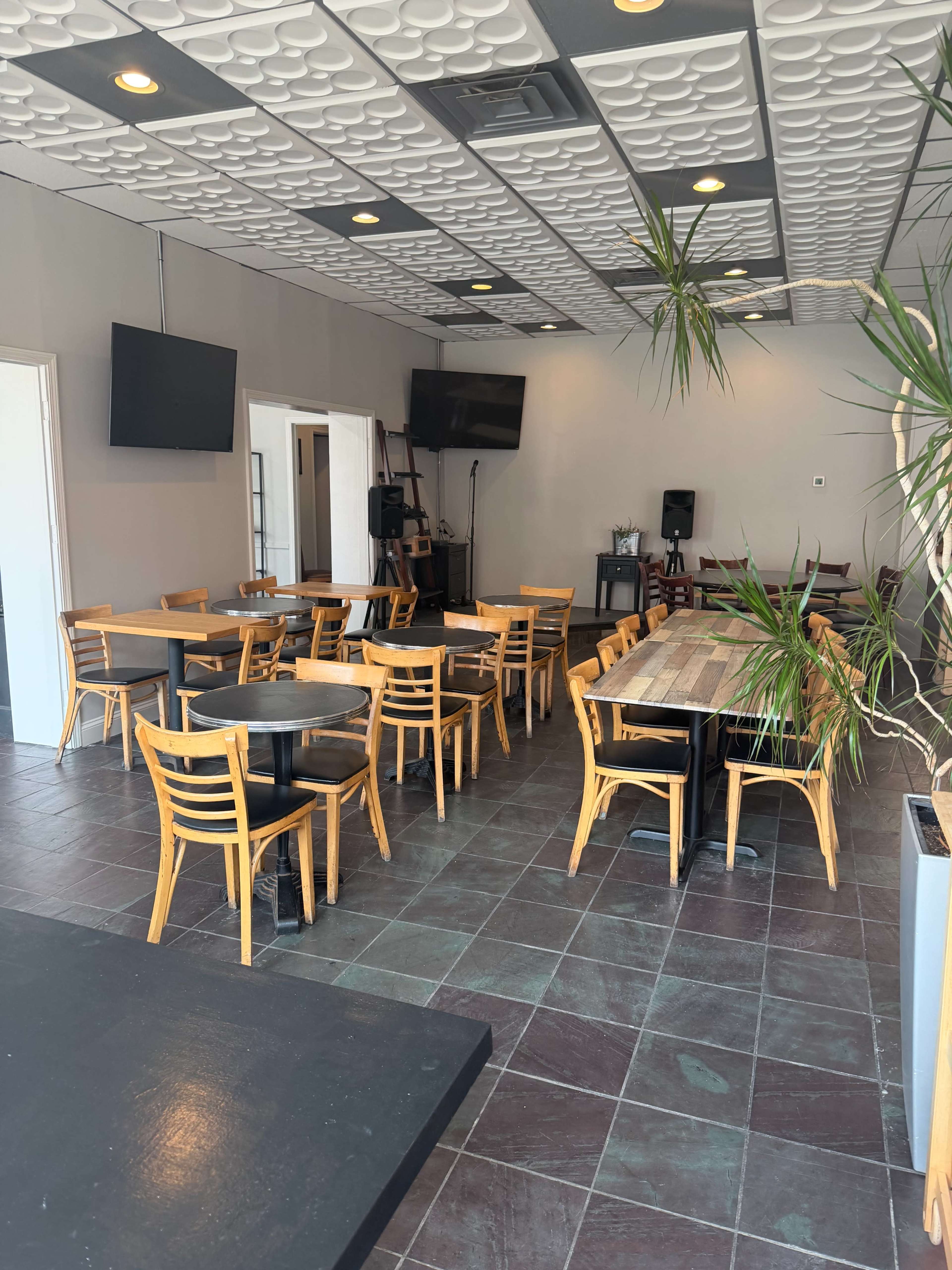 The image shows an empty restaurant interior with wooden tables and chairs arranged in a simple layout, featuring hanging plants and screens mounted on the walls.
