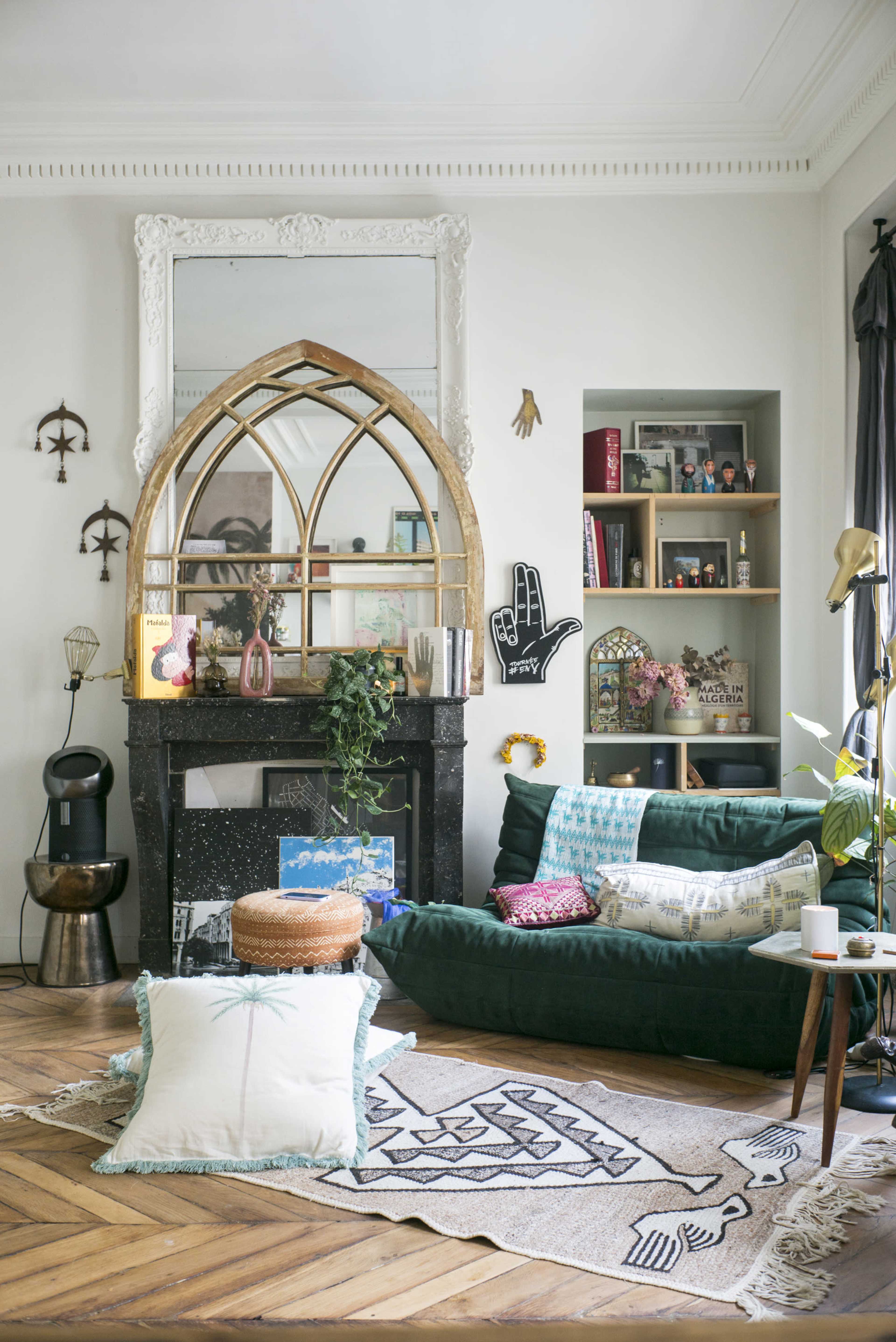 A cozy living room features a dark green sofa, a large arched mirror above a black fireplace, and a patterned rug on a wooden floor, accented by decorative shelves and plants.