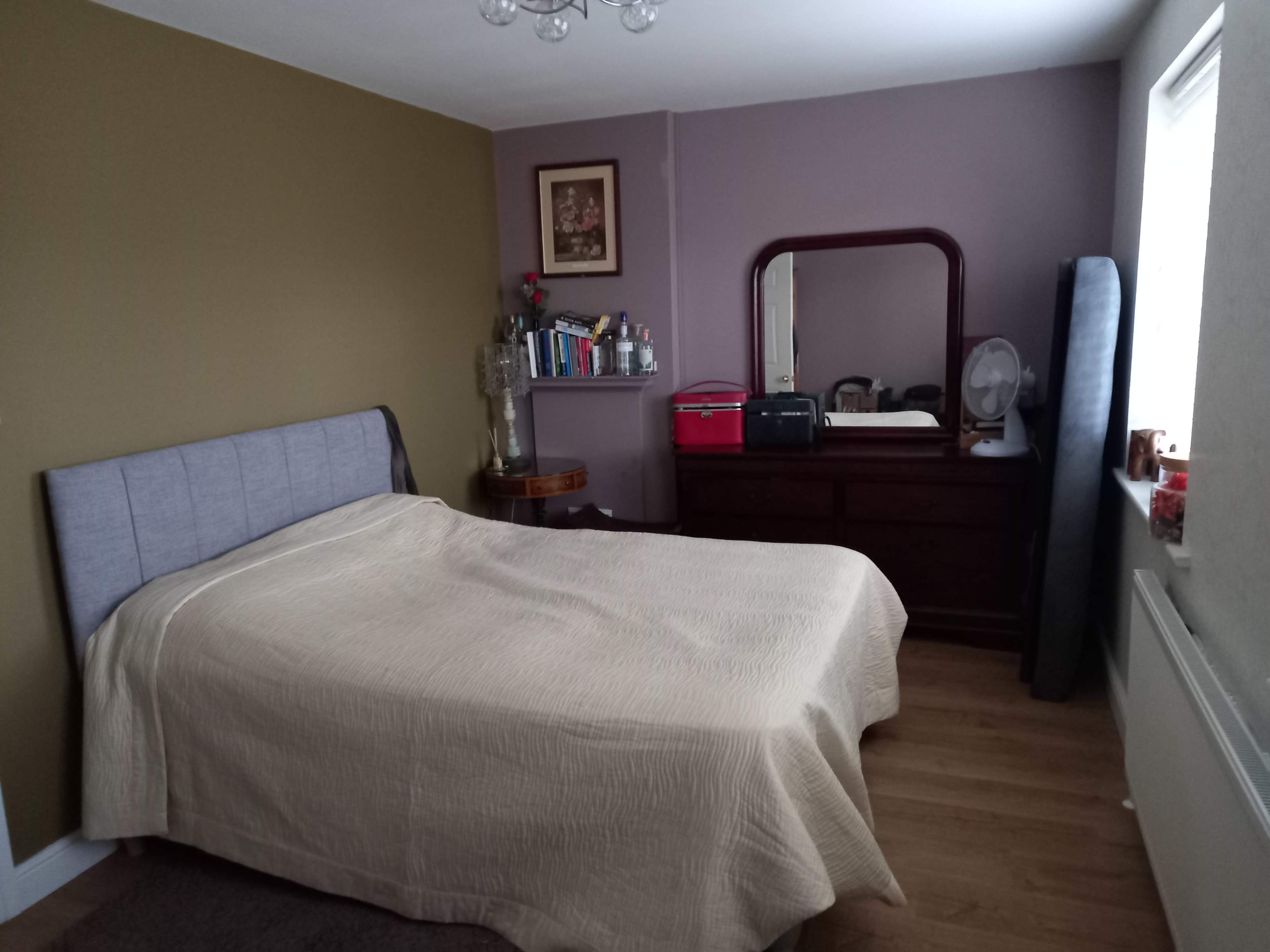 A simple bedroom with a bed covered by a light beige blanket, a wooden dresser with a mirror, books on a shelf, and a window with natural light.